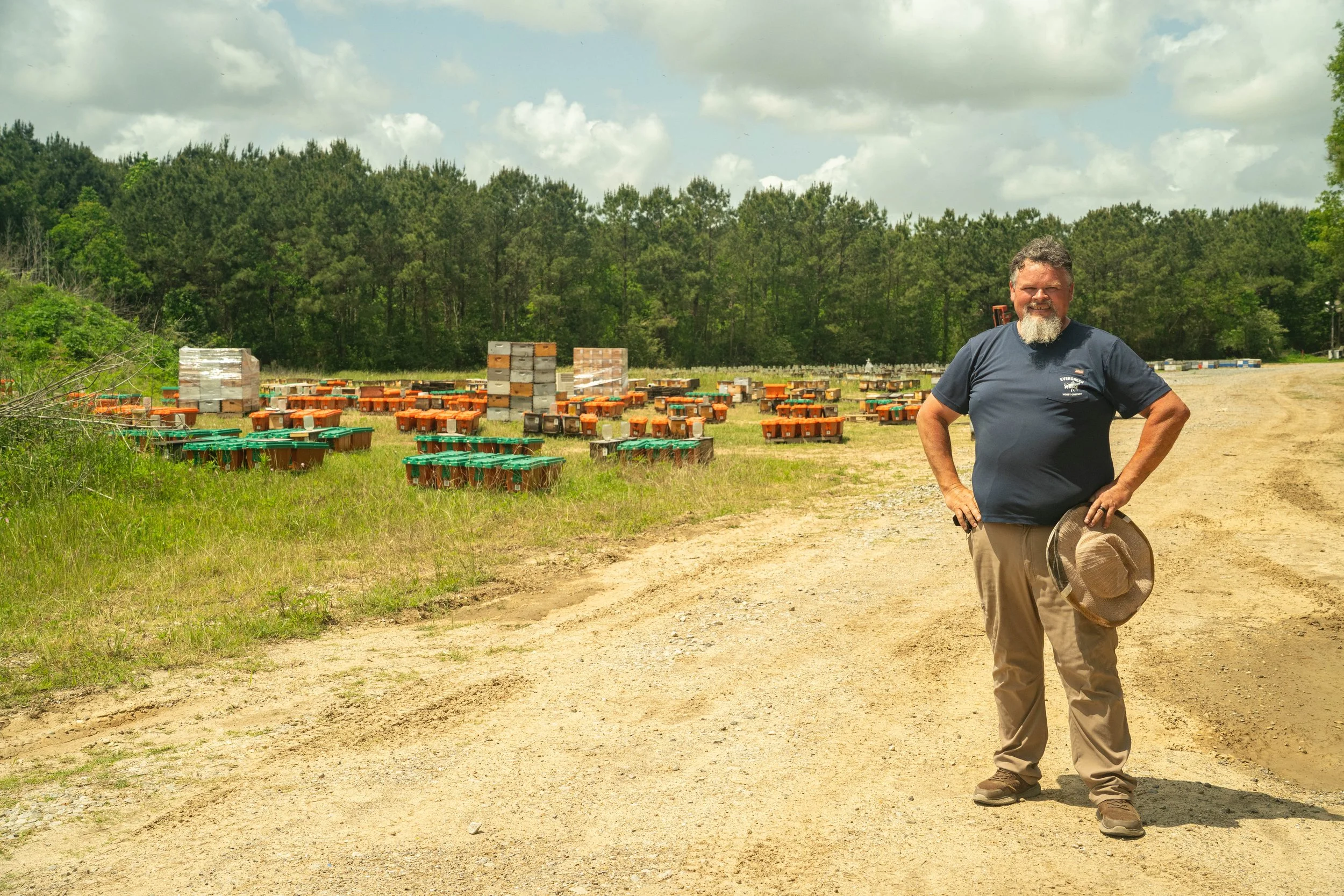 Wes Card standing on a dirt path in Louisiana, smiling, holding his bee keeper hat, with numerous bee boxes behind him, surrounded by green trees under a partly cloudy sky.