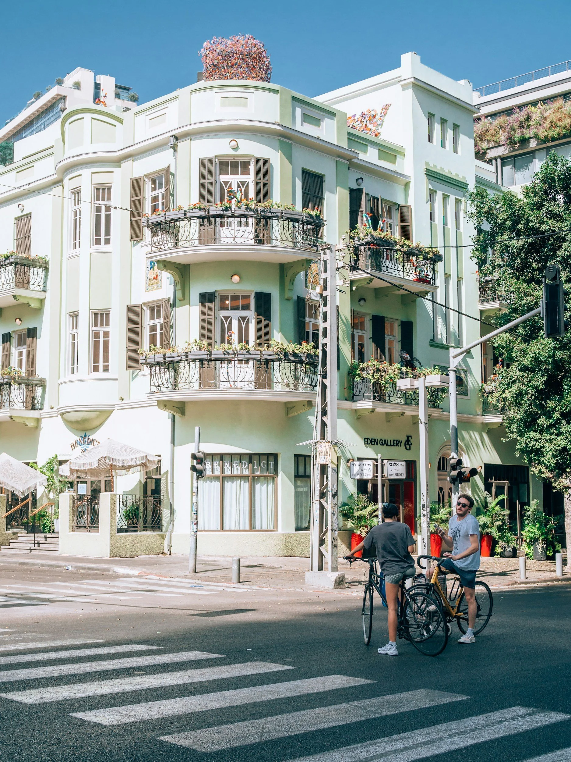 Street scene with two men on bicycles next to a pedestrian crosswalk, in front of a pastel green building with balconies and flower boxes, a tree, and a traffic signal.