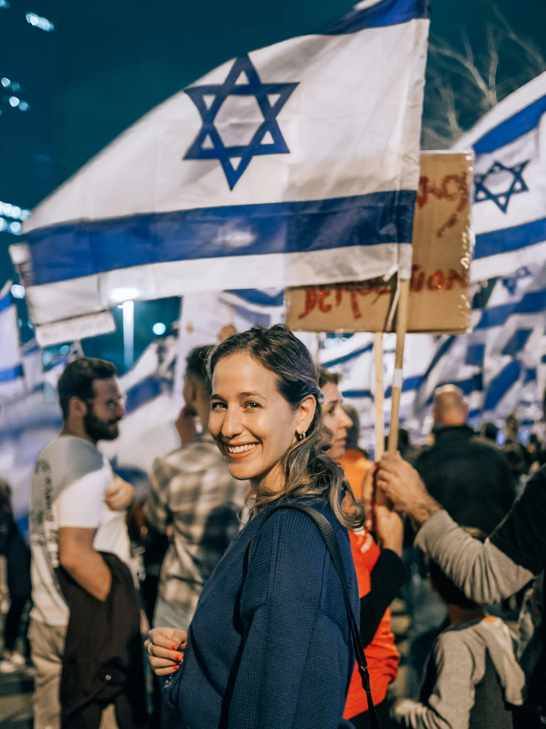 A woman with long hair smiles at the camera at a gathering or demonstration, surrounded by other people, with Israeli flags in the background.