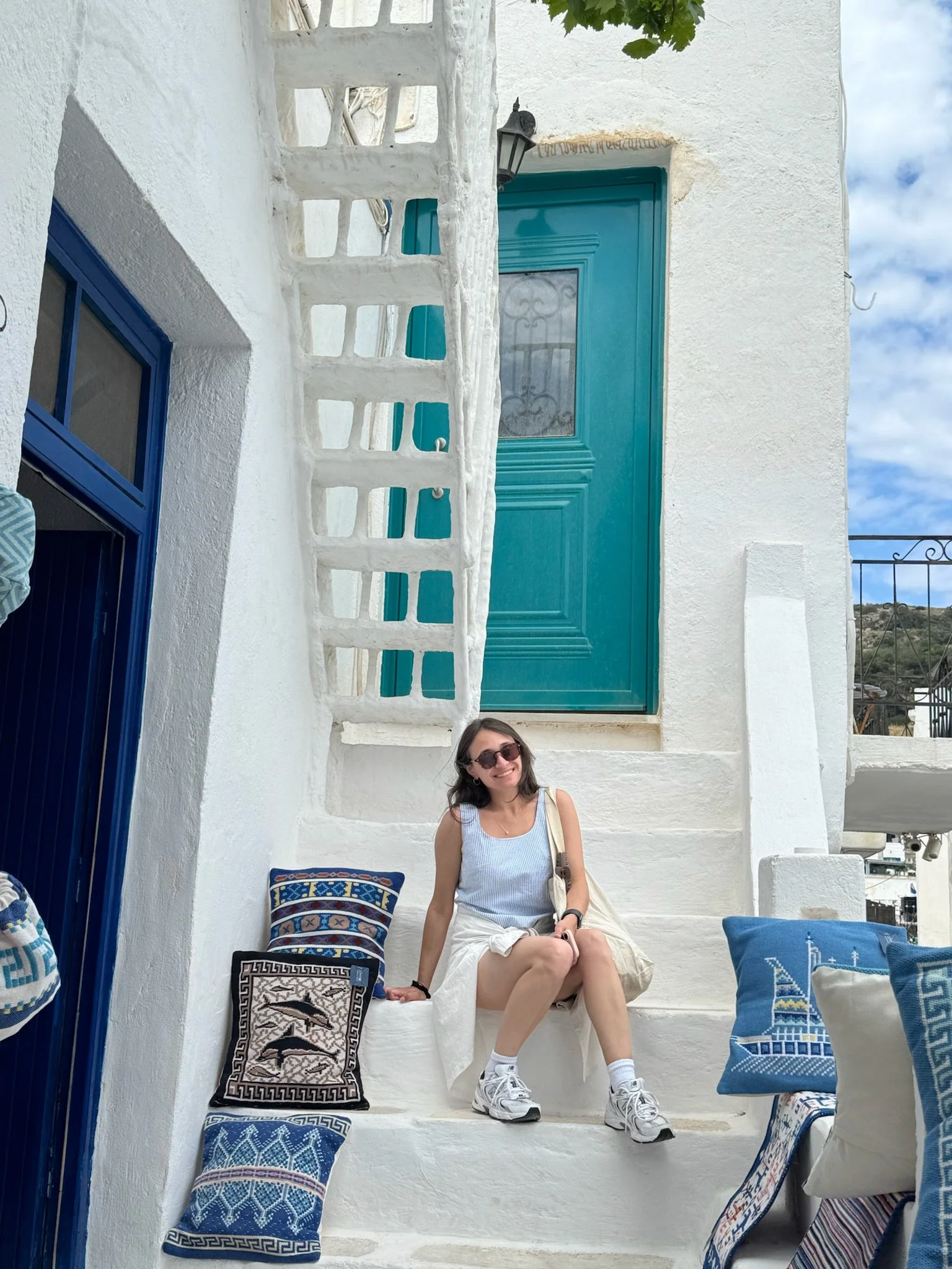Smiling young woman with sunglasses sitting on a white stair in front of a white building with blue doors and windows. Decorated cushions with Greek patterns and blue and white colors are on either side of her. The building has a small outdoor light and a balcony railing visible on the right side. Part of the sky and some distant landscape are visible in the background.