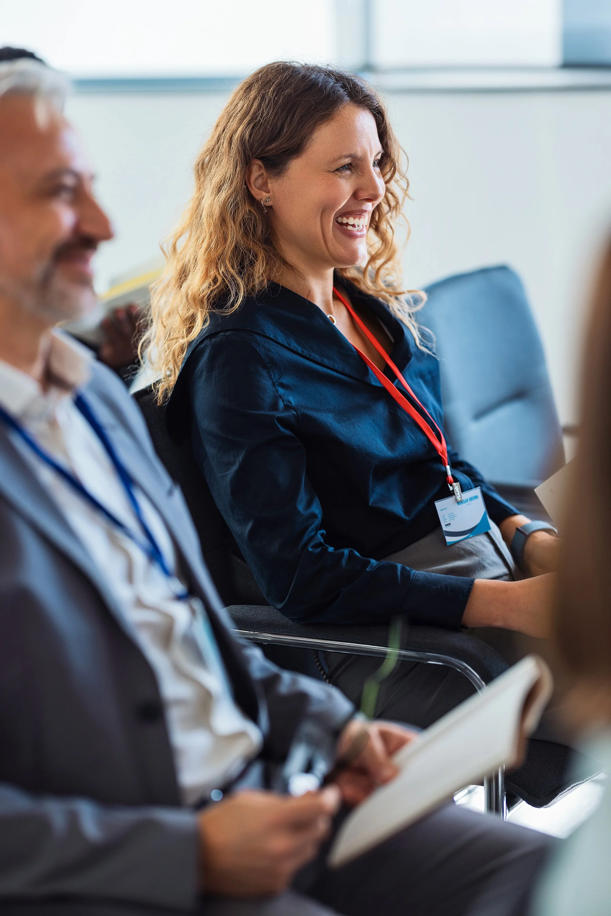 A woman with curly brown hair smiling during a meeting, seated next to men, holding a tablet, in a modern conference room.