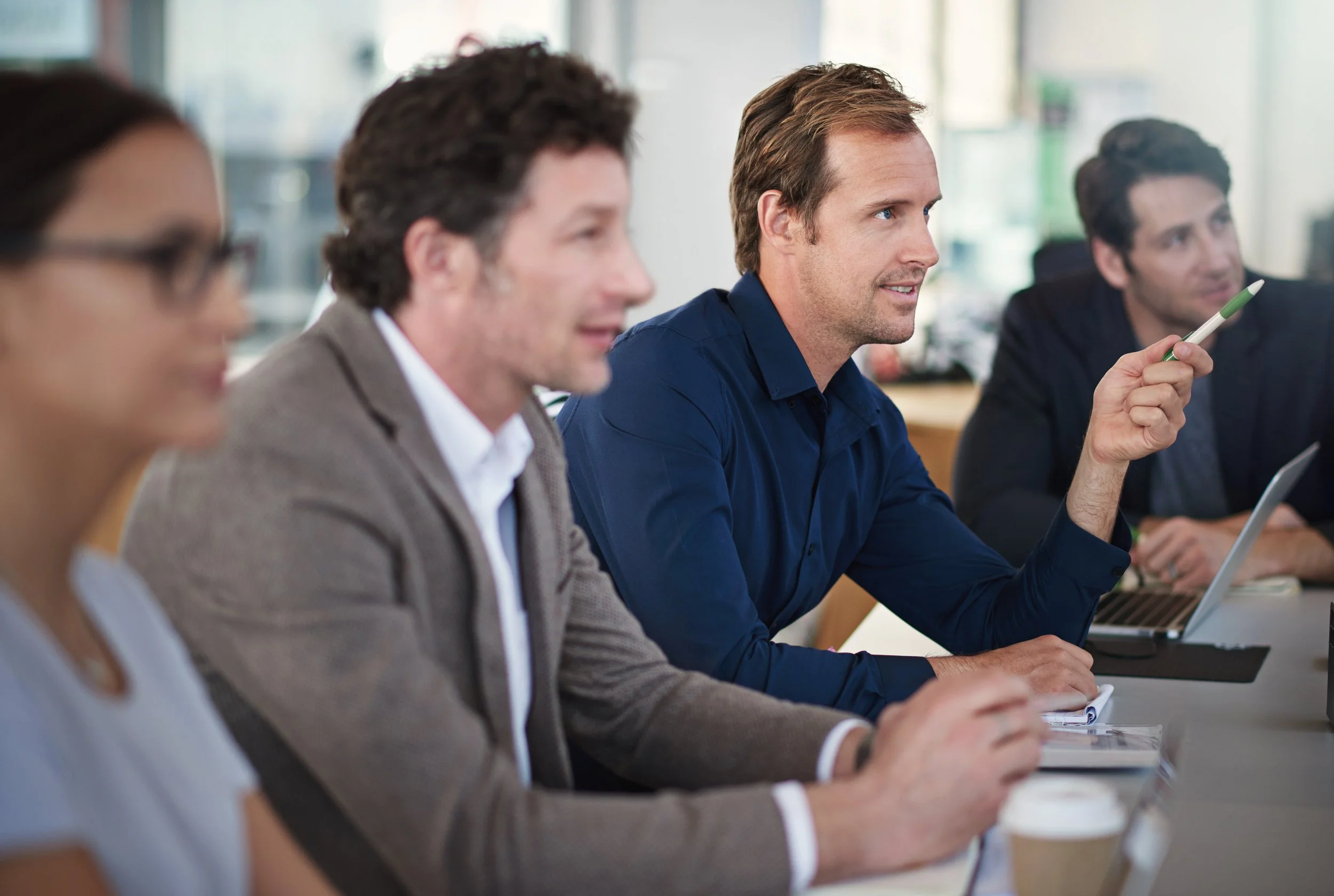 A group of professionals sitting at a conference table, listening attentively during a meeting. The man in the blue shirt is holding a pen.