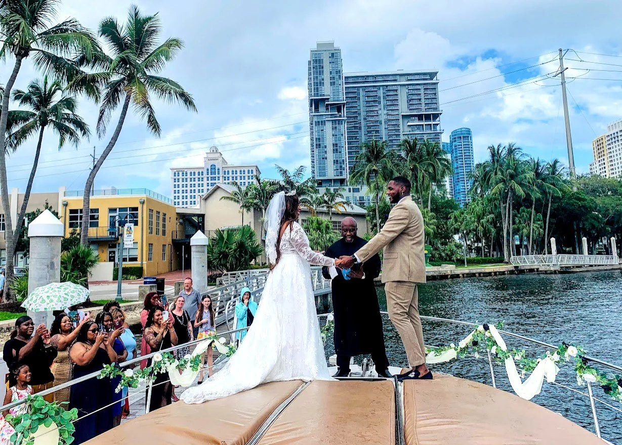 A couple getting married on a boat by the water, with guests watching and taking photos. The bride is wearing a white wedding dress, and the groom is in a beige suit. The ceremony is outdoors in an urban area with palm trees, high-rise buildings, and some colorful houses.