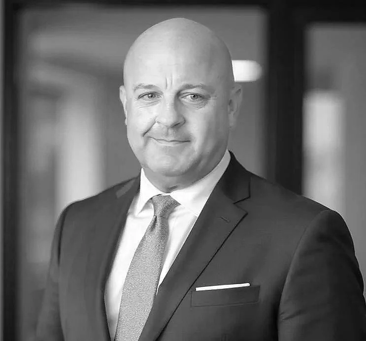 Black and white photo of a man in a suit and tie, smiling slightly with a neutral background.