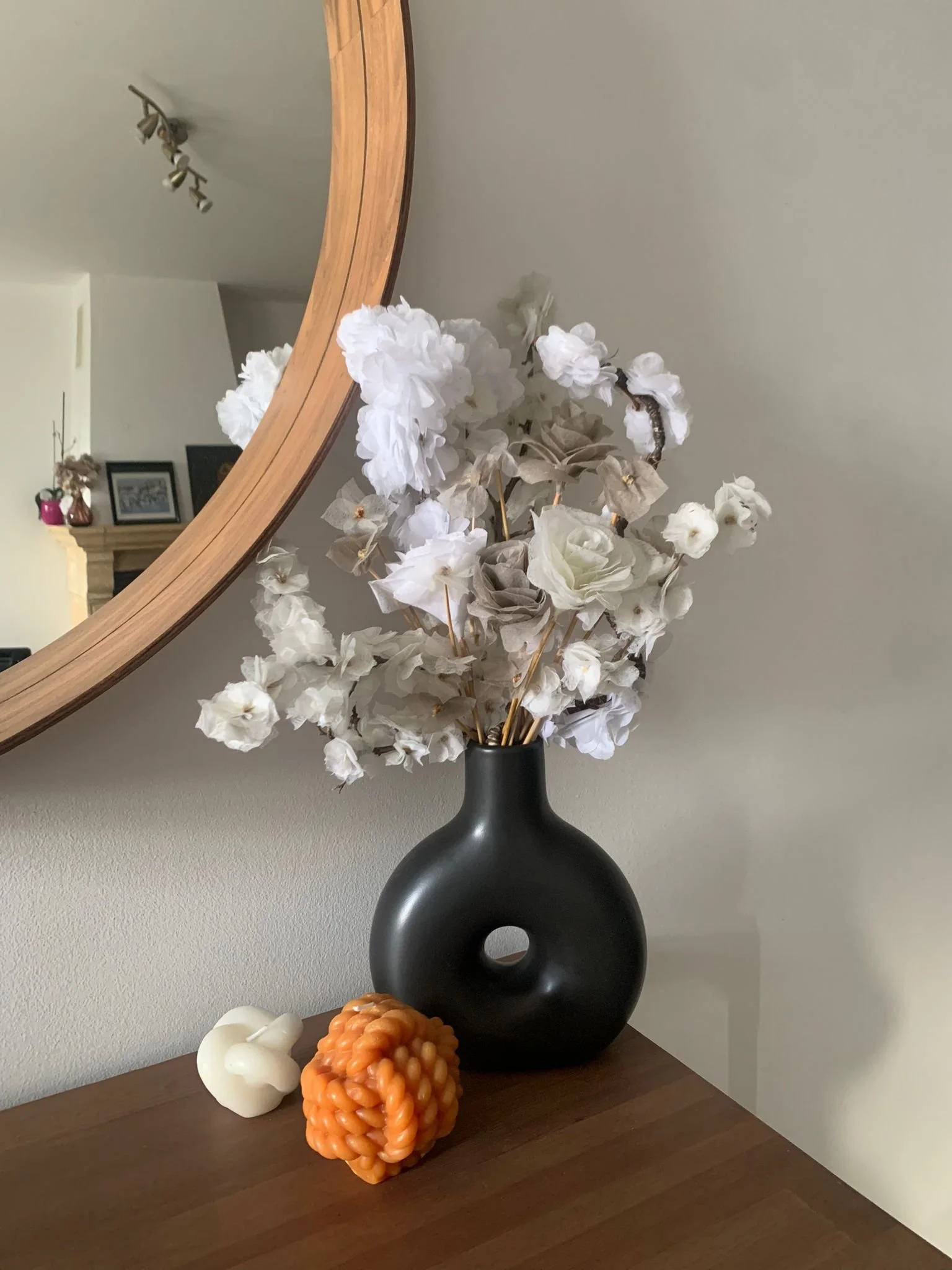 Un vase noir avec des fleurs blanches artificielles, un décor avec une boule orange en caoutchouc, une sculpture blanche en forme de cœur et une pièce orange sous un miroir rond en bois sur une table en bois massif.
