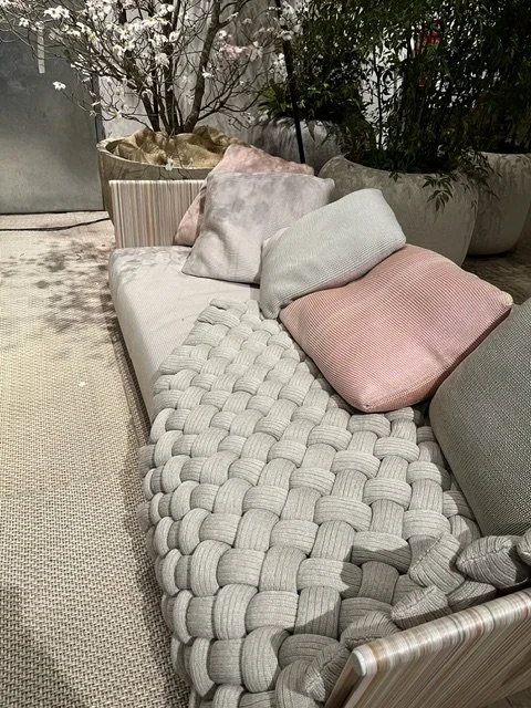 Ein Gartenmobiliar mit einem gepolsterten Sitzkissen und mehreren zusammengerollten Kissen in Pastellfarben, umgeben von Pflanzen und Blumentöpfen.
