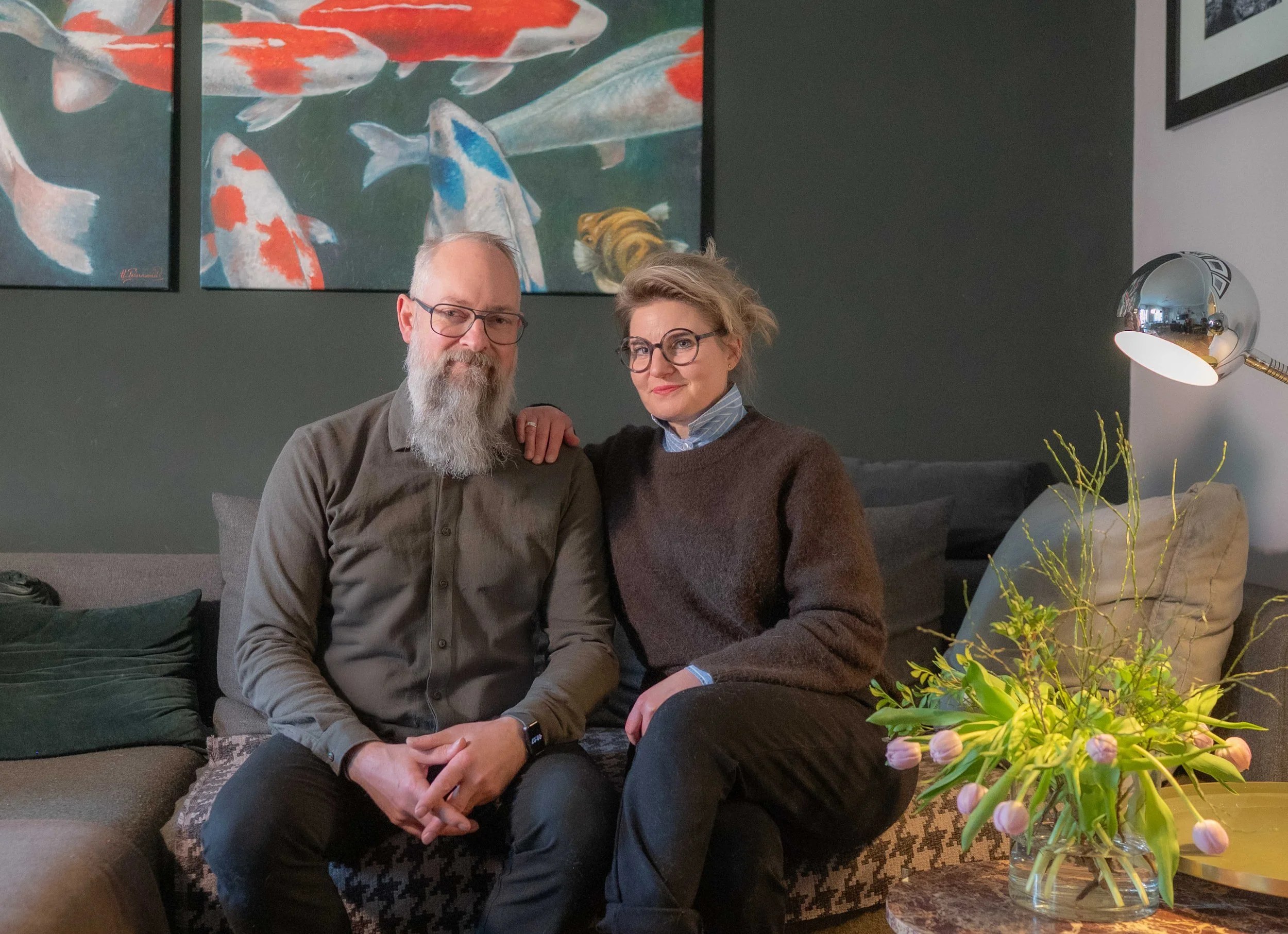 Ein Mann mit Brille, Bart und grauem Haar sowie eine Frau mit Brille und gewelltem Haar sitzen auf einer Couch in einem Wohnzimmer und schauen in die Kamera. Im Hintergrund sind Bilder von Koi-Fischen an der Wand, und es gibt einen Tisch mit einem Blumenstrauß auf der rechten Seite.