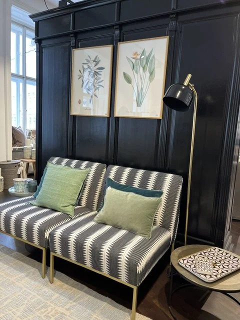 Ein Wohnzimmer mit schwarz lackierten Wänden, einer schwarzen und weißen gestreiften Couch, zwei grünen Kissen, einer Stehlampe und zwei makellosen botanischen Wandbildern.