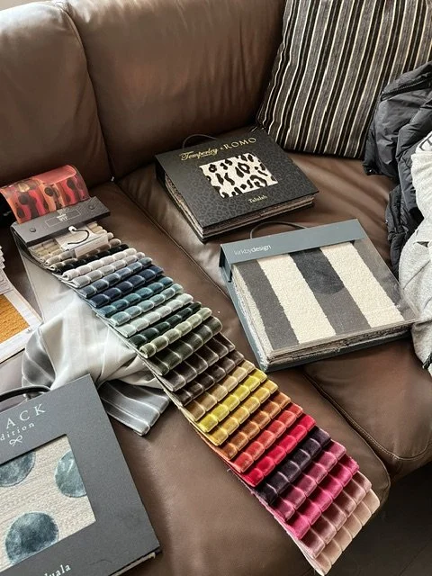 Stoffmuster und Farbproben auf einem braunen Sofa, inklusive Garn- und Musterbüchern.