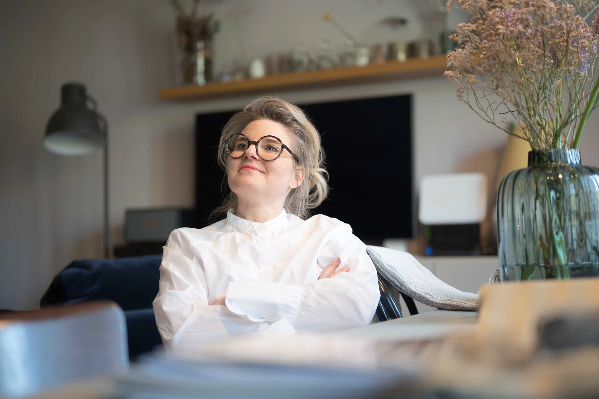 Nicole mit Brille, die lächelnd im Wohnzimmer sitzt, umgeben von Dekorationen und Pflanzen.