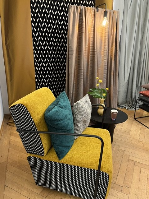 Gemütliches Wohnzimmer mit gelbem Sofa, bunten Kissen, einem schwarzen Beistelltisch mit Vase und Blume, Vorhängen vor einem Fenster, Holzboden.