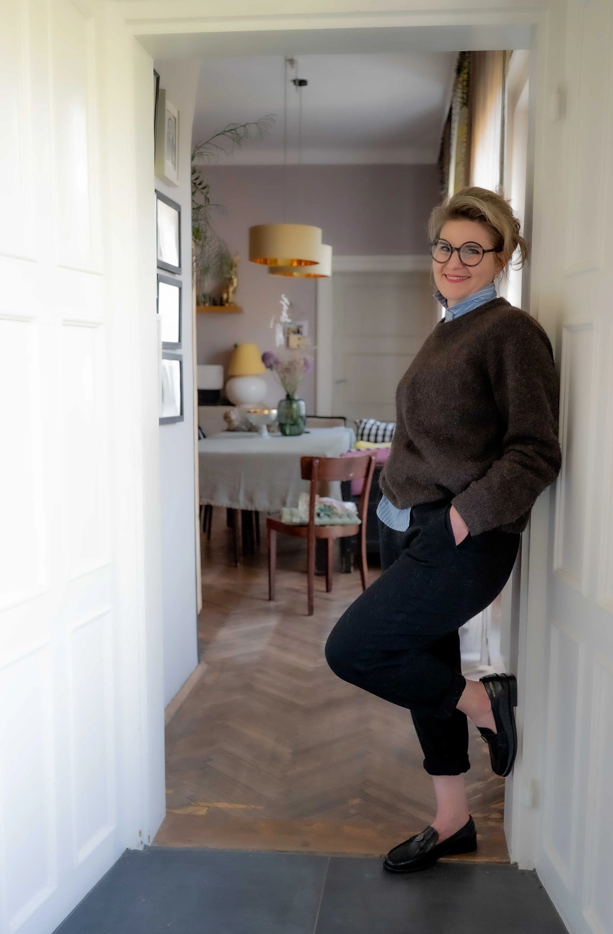 Eine Frau, Nicole, lächelt in einem Wohnzimmer, lehnt an der Türrahmen, trägt schwarze Schuhe, eine braune Pullover, Brille und hat die Hände in den Taschen.