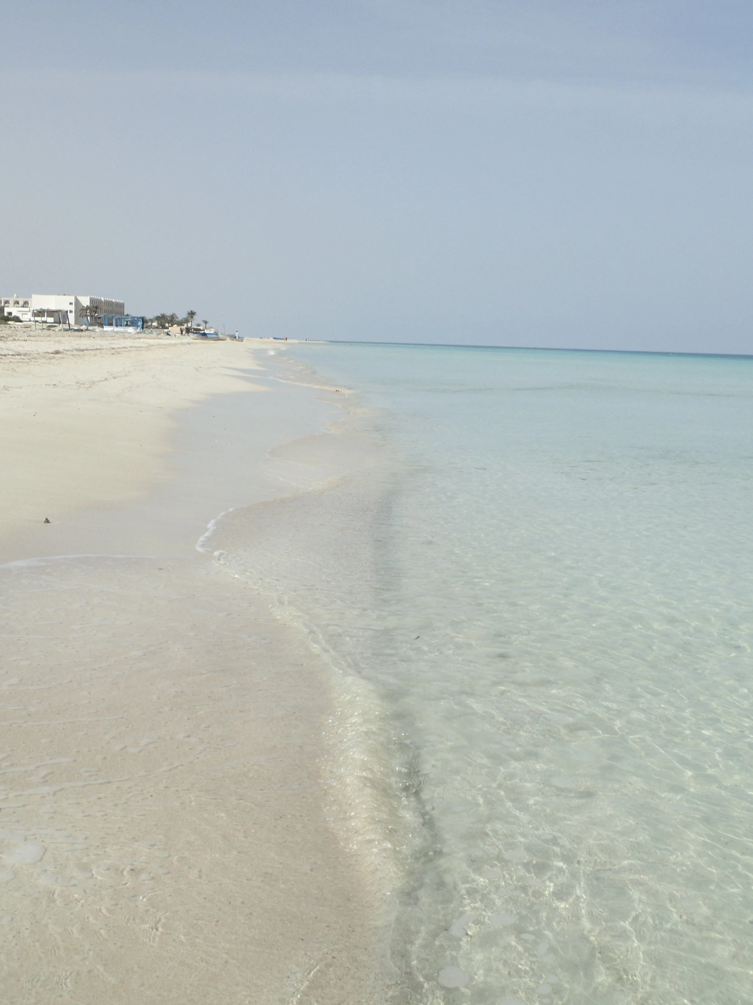 Clear, calm ocean water washing onto a white sandy beach with distant buildings and palm trees under a slightly cloudy sky