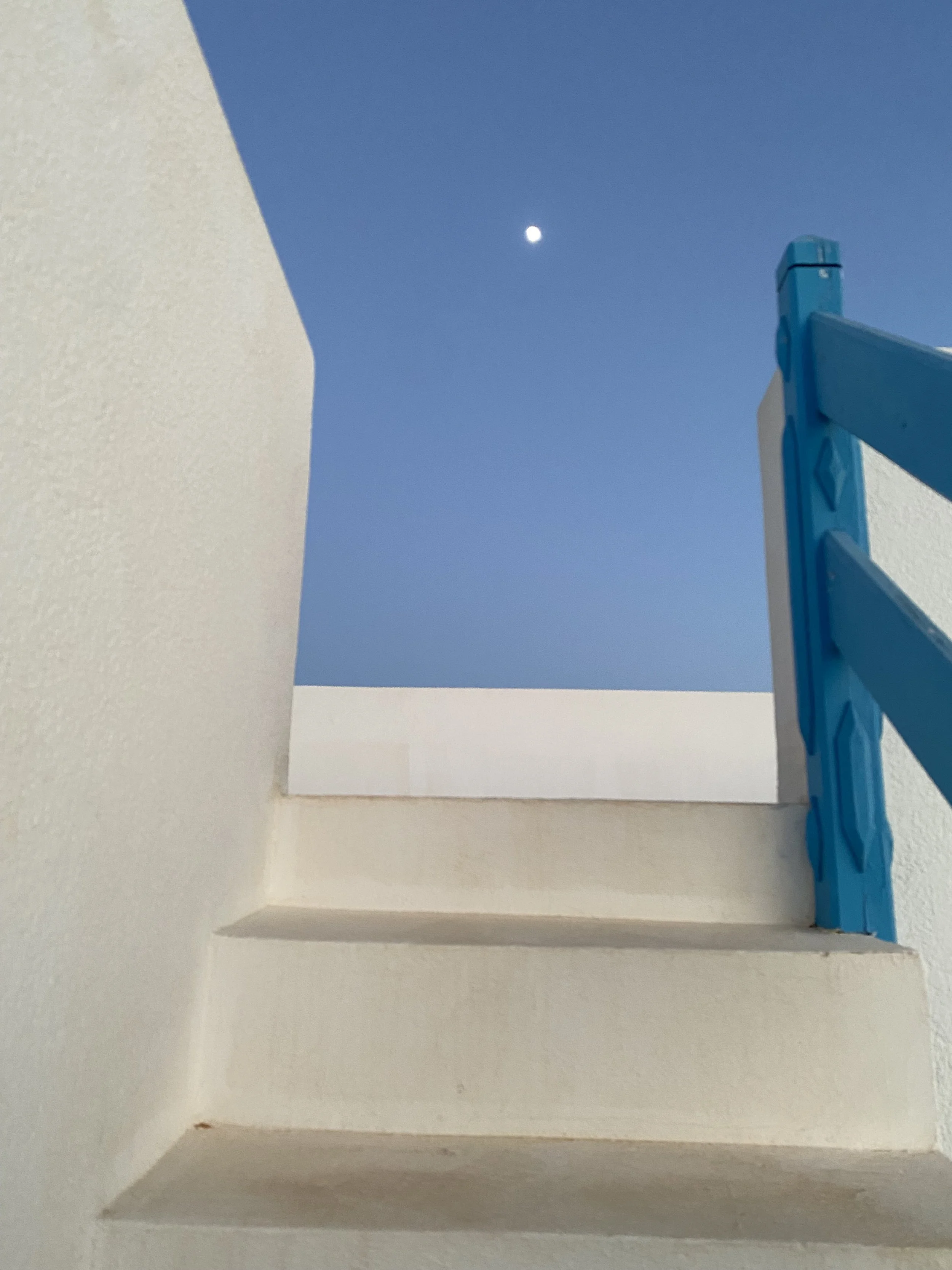 A view of a blue sky with a full moon, partially visible through a small window or opening in a wall, with a blue and white staircase on the right side of the image.