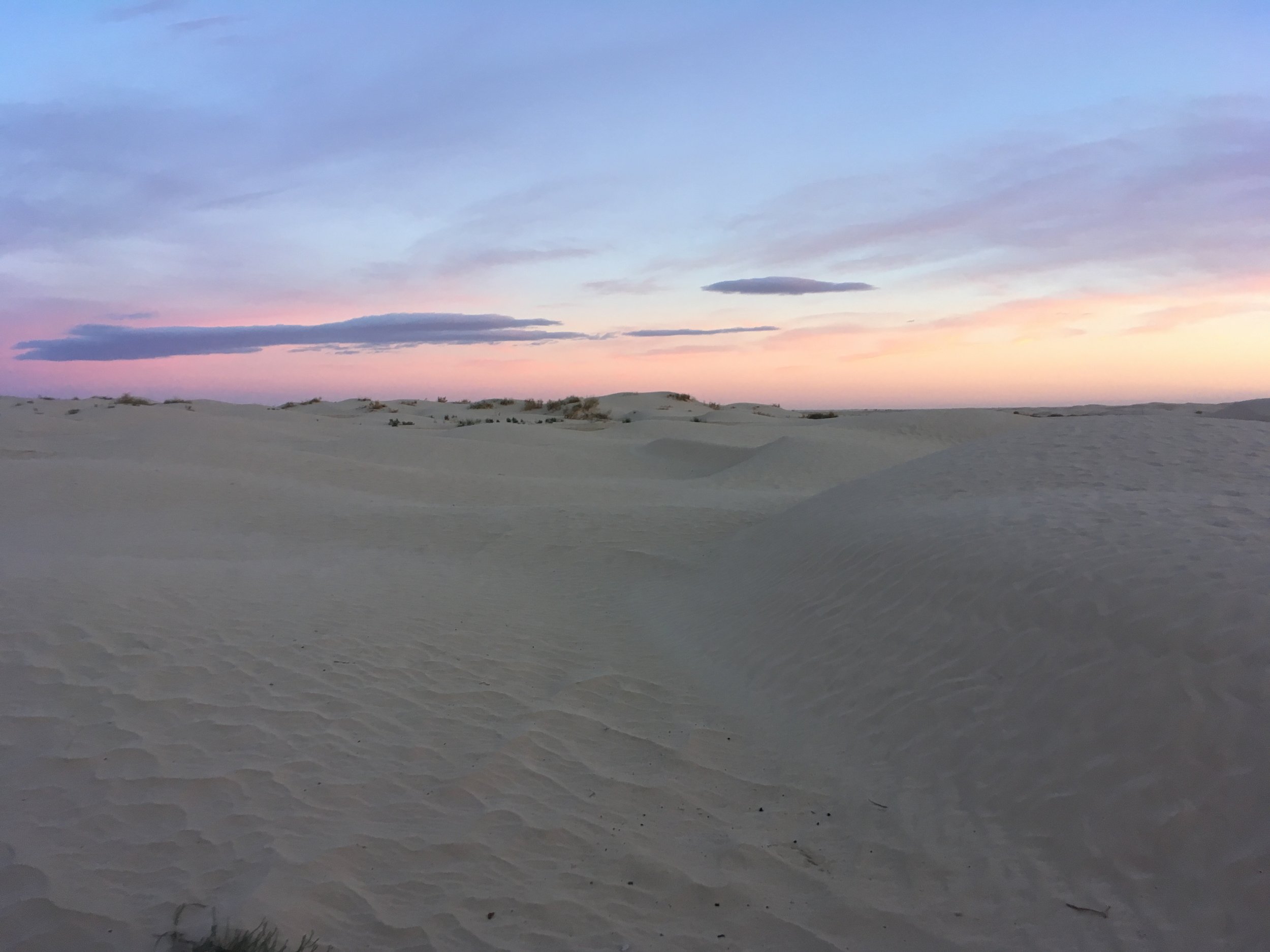 A landscape of sand dunes under a colorful sky with pink, purple, and blue hues, and some scattered clouds.