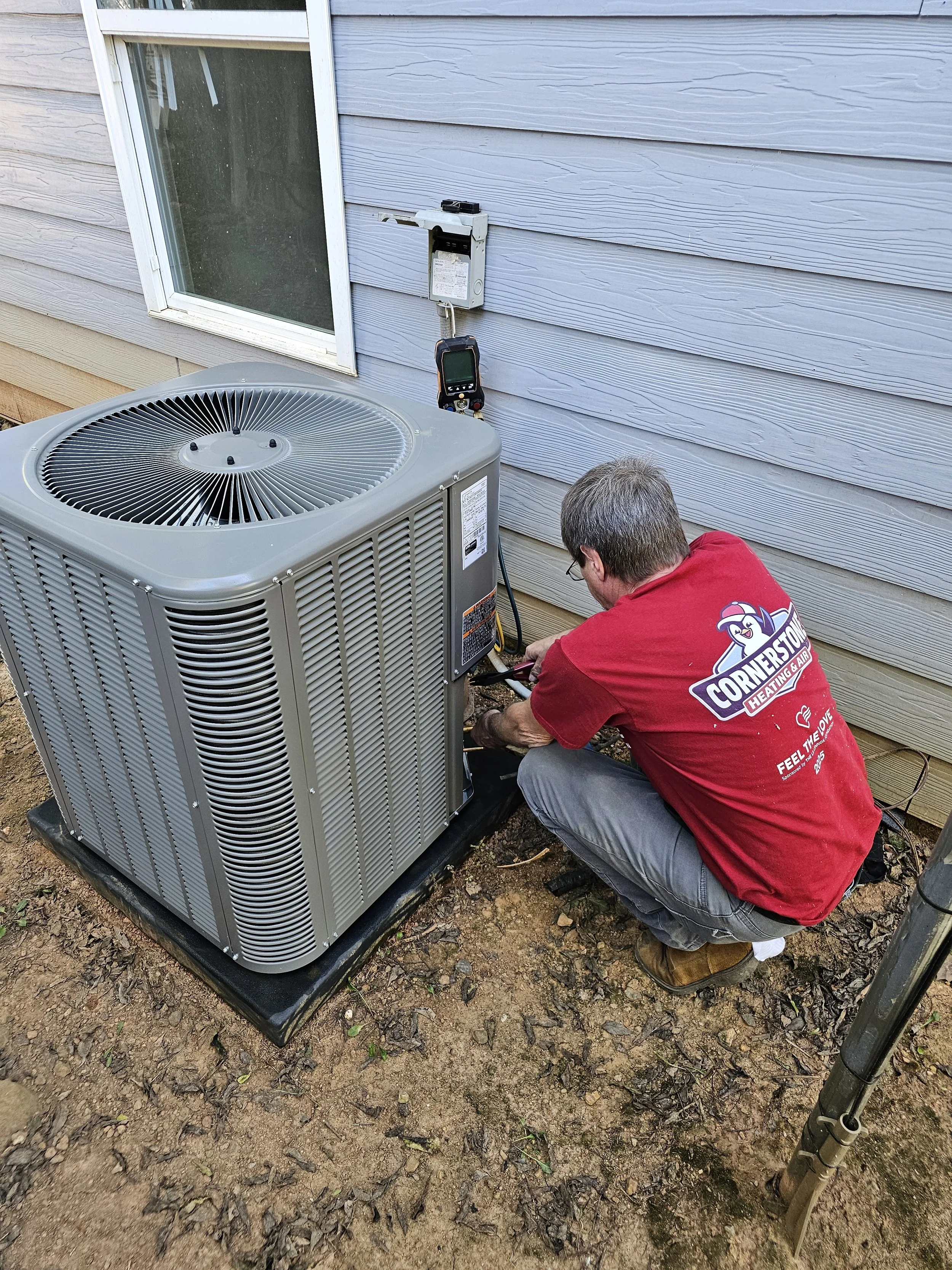 A man in a red shirt working on an outdoor air conditioning unit, crouched down and using tools, with a gray house siding and window in the background.
