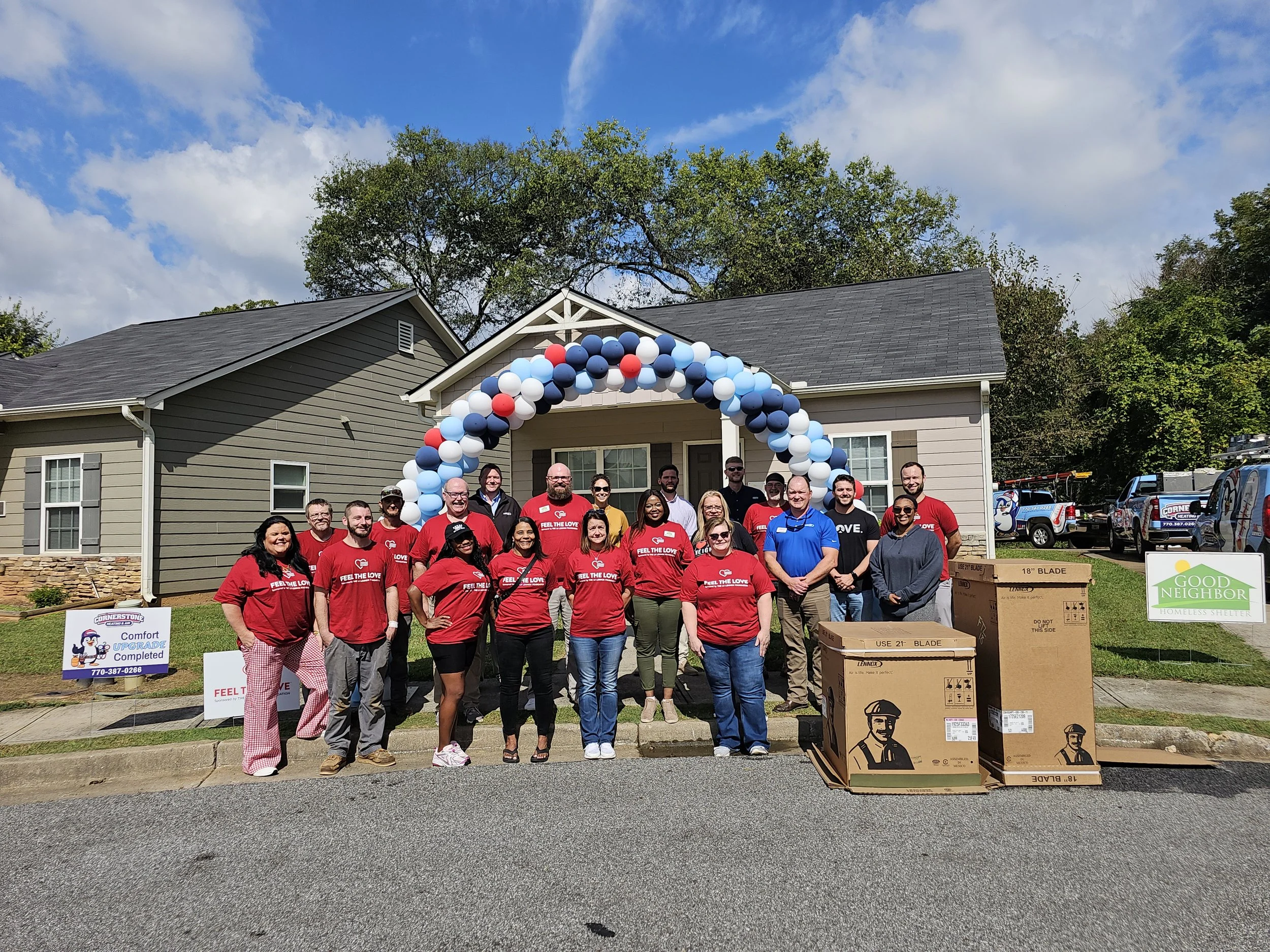 A group of people standing in front of a house with a balloon arch and donation signs, participating in a community event to support homelessness. There are boxes and trucks nearby.