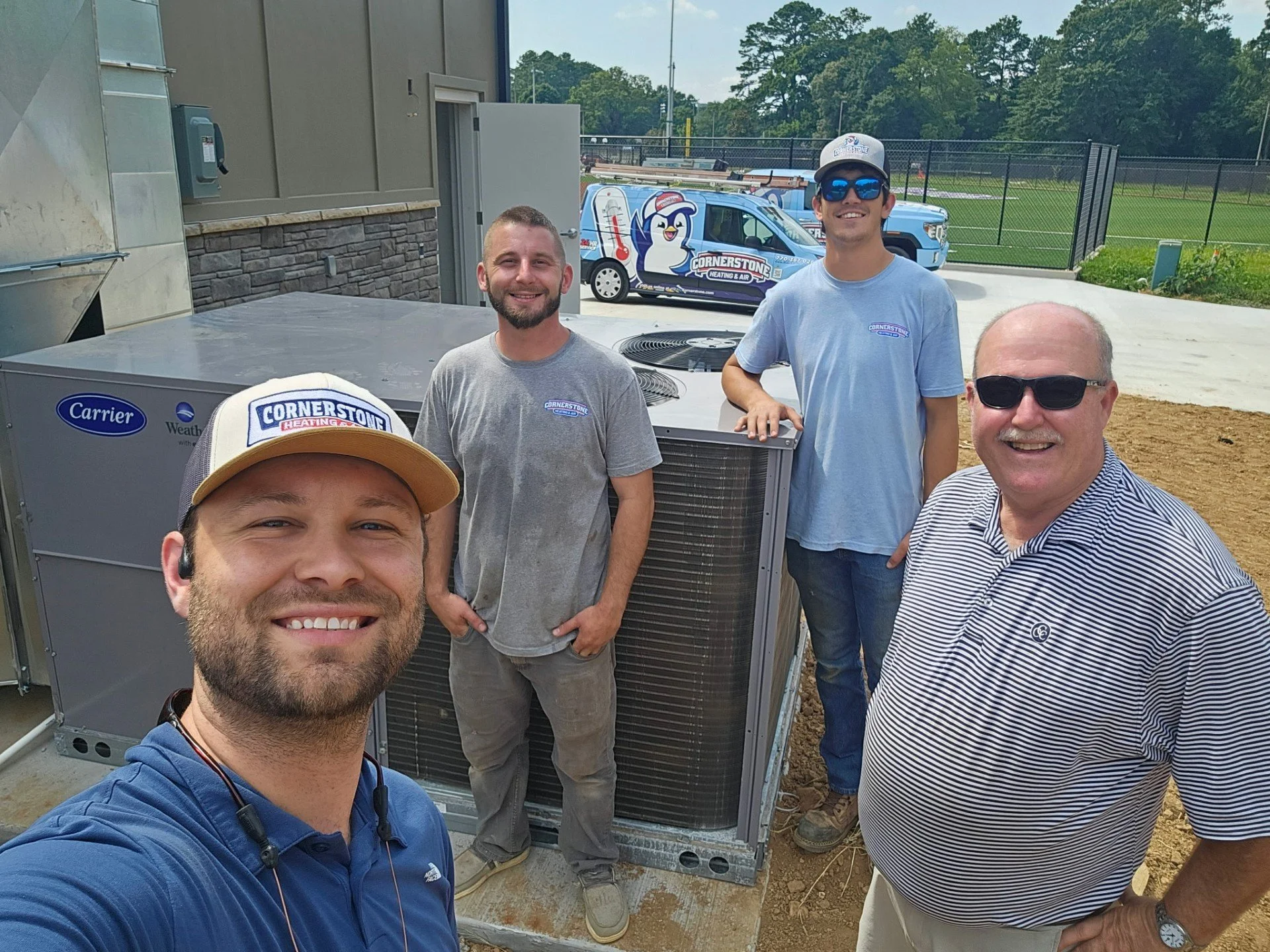 Group of five men smiling outdoors near HVAC equipment, with a sports field in the background, some wearing hats and sunglasses.