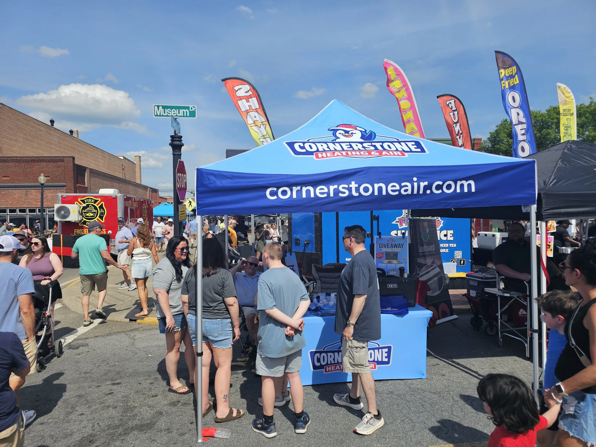 A busy outdoor street fair with a blue tent for Cornerstone Heating & Air, surrounded by many people walking and shopping, with colorful flags and a clear blue sky overhead.
