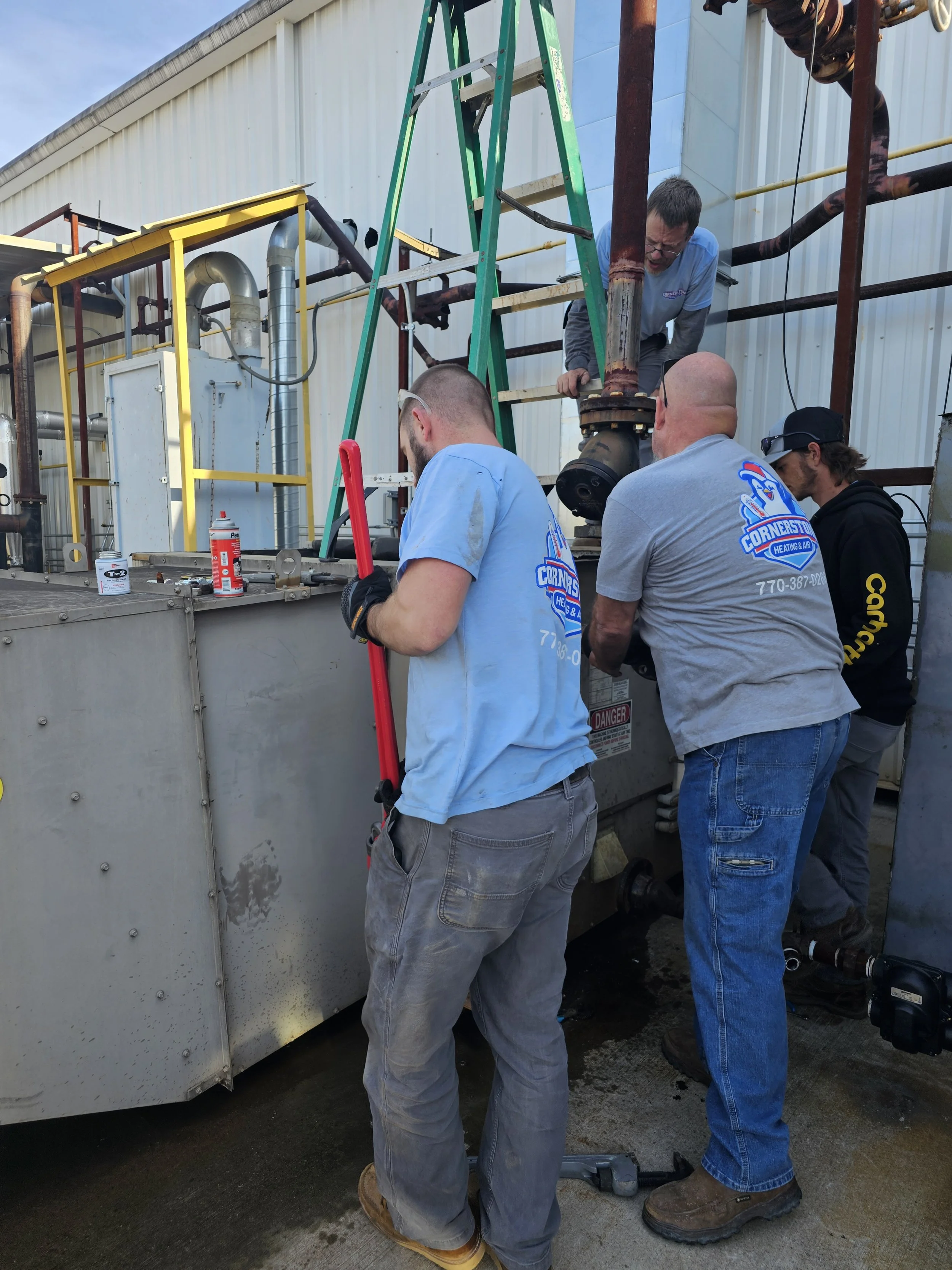 Four workers repairing or inspecting equipment outside a large industrial unit. They are using tools and some are wearing gloves and safety glasses. The setting is outdoors near the side of a building with pipes and metal siding.
