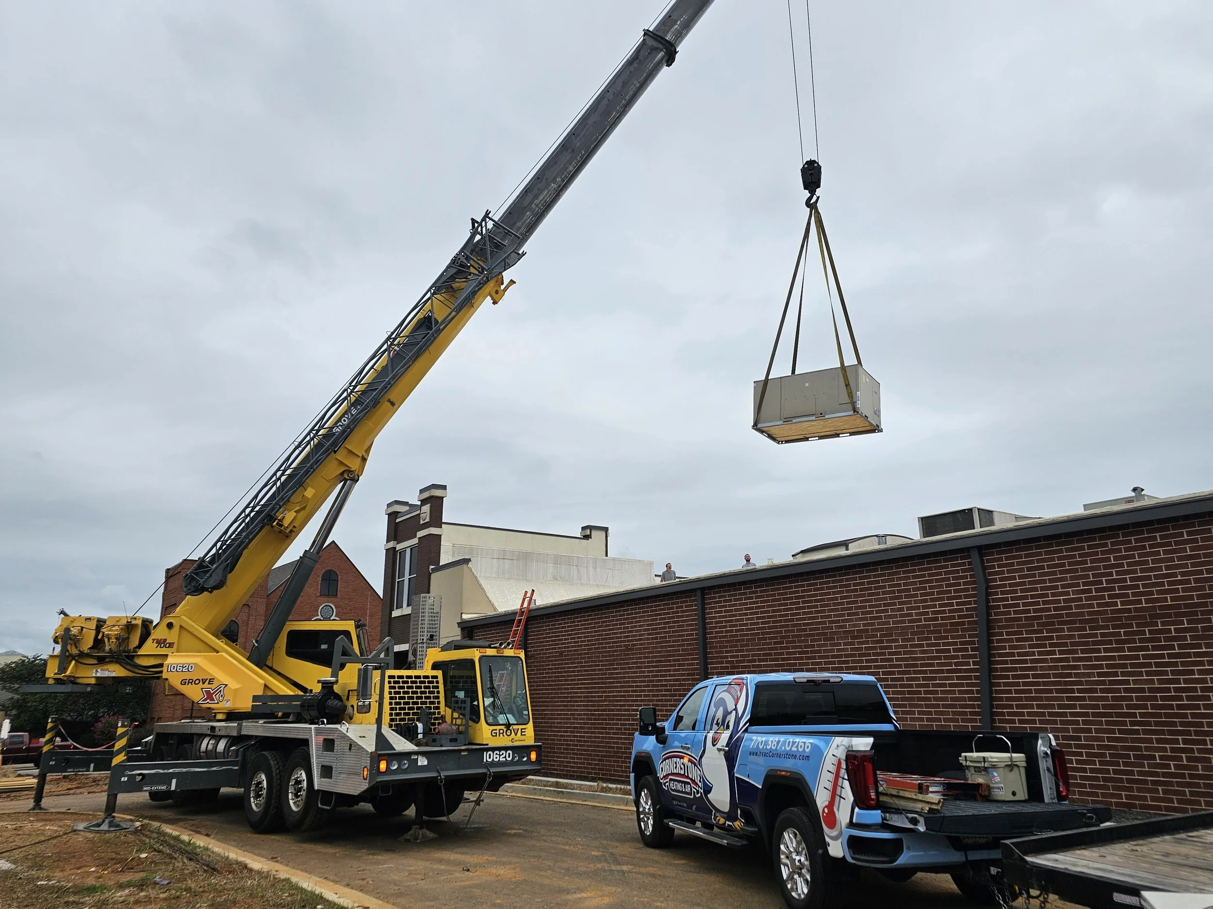 A yellow crane lifting a large box on a pallet, with a blue van and a decorated blue vehicle parked nearby, in front of a brick building under cloudy sky.