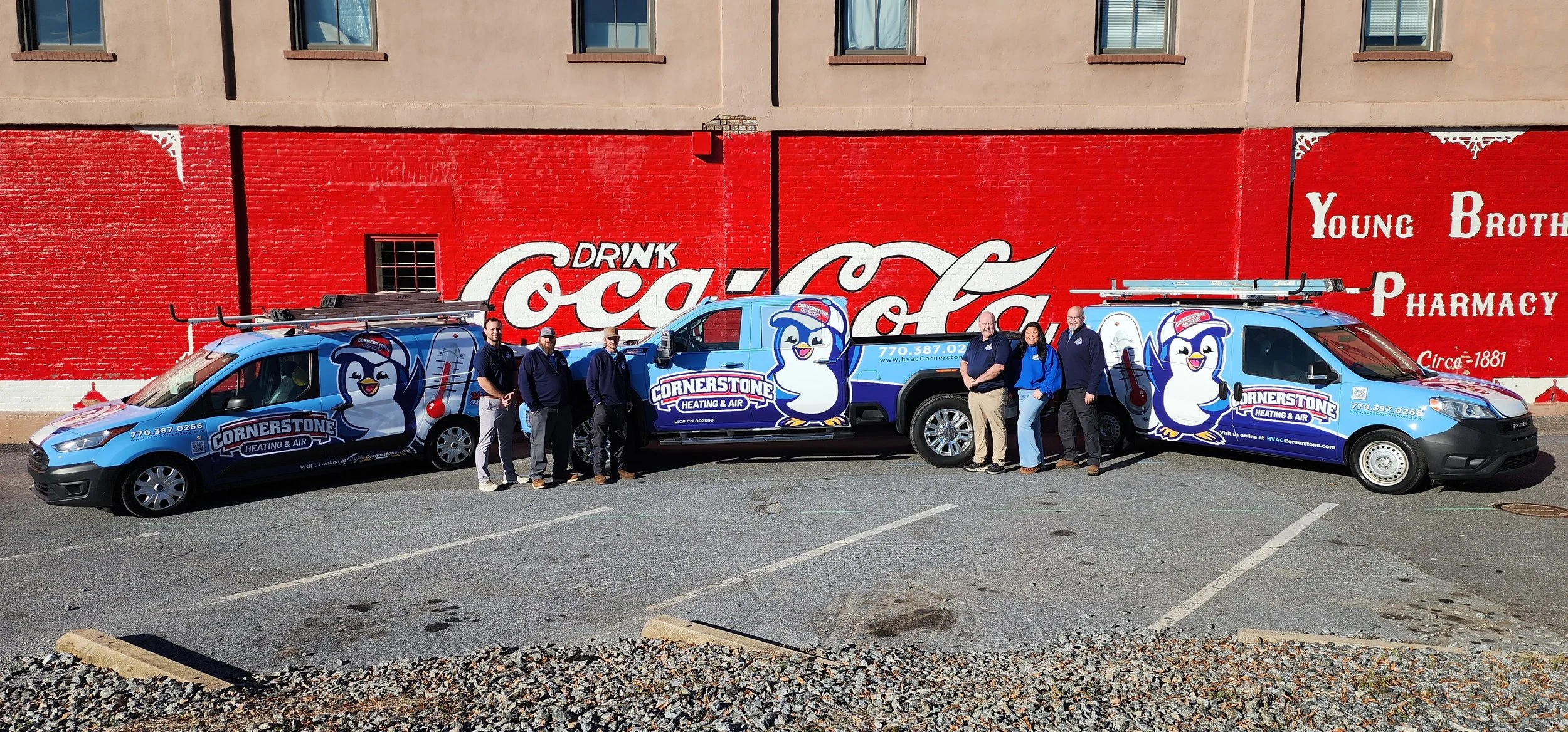 A group of six people standing in front of two service vehicles branded with the Cornerstone Heating & Air logo, parked in front of a red wall with a large Coca-Cola mural.