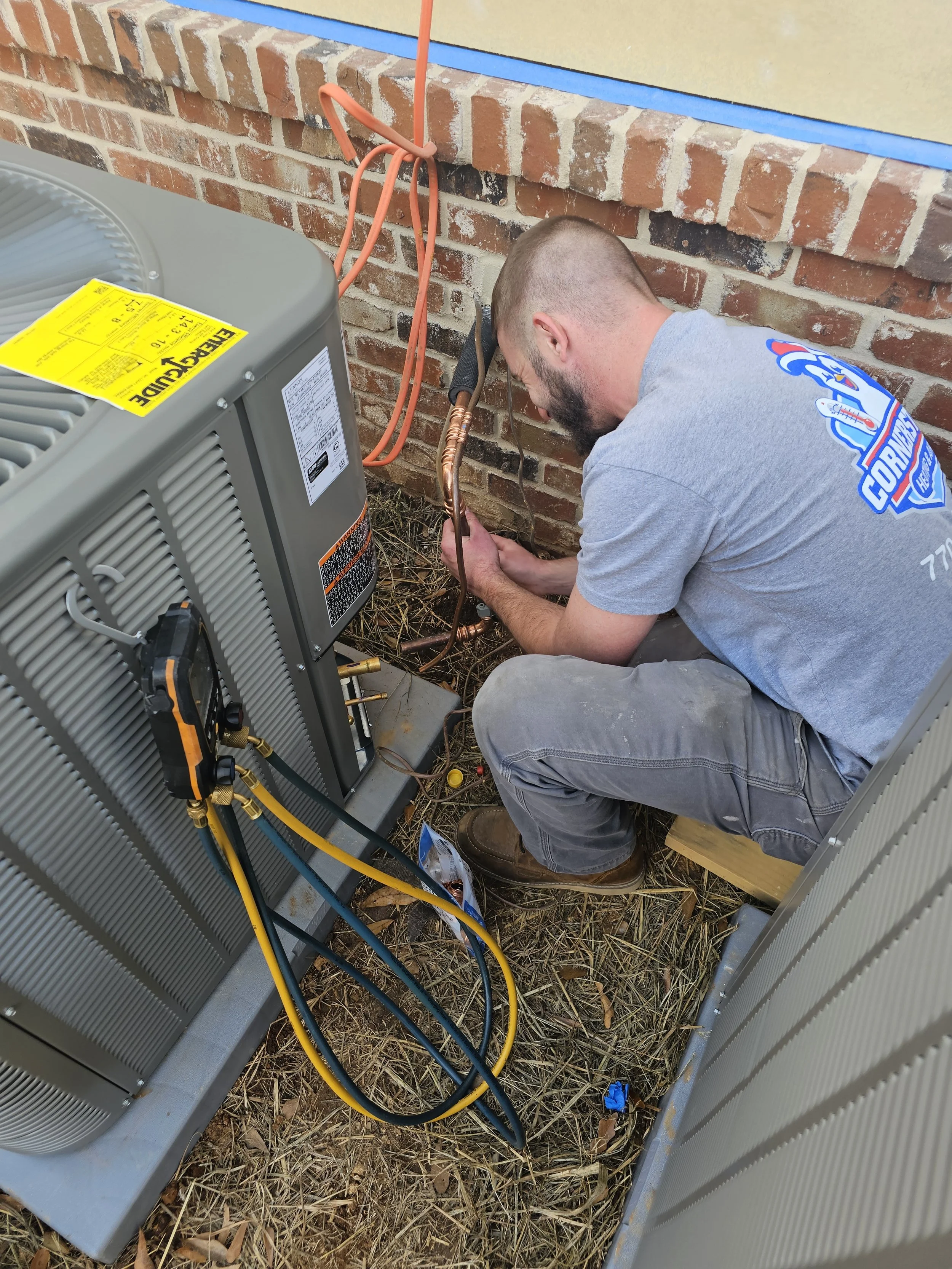 A technician crouched outside working on an HVAC unit, using tools to connect copper pipes, with a brick wall behind him and some electrical wires in the scene.