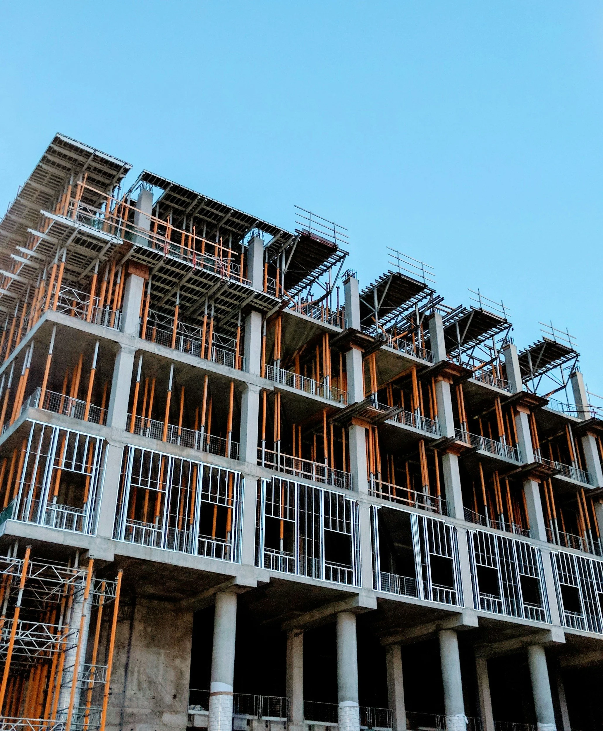 Construction site with a multi-story apartment building under construction, scaffolding, and support beams, against a clear blue sky.
