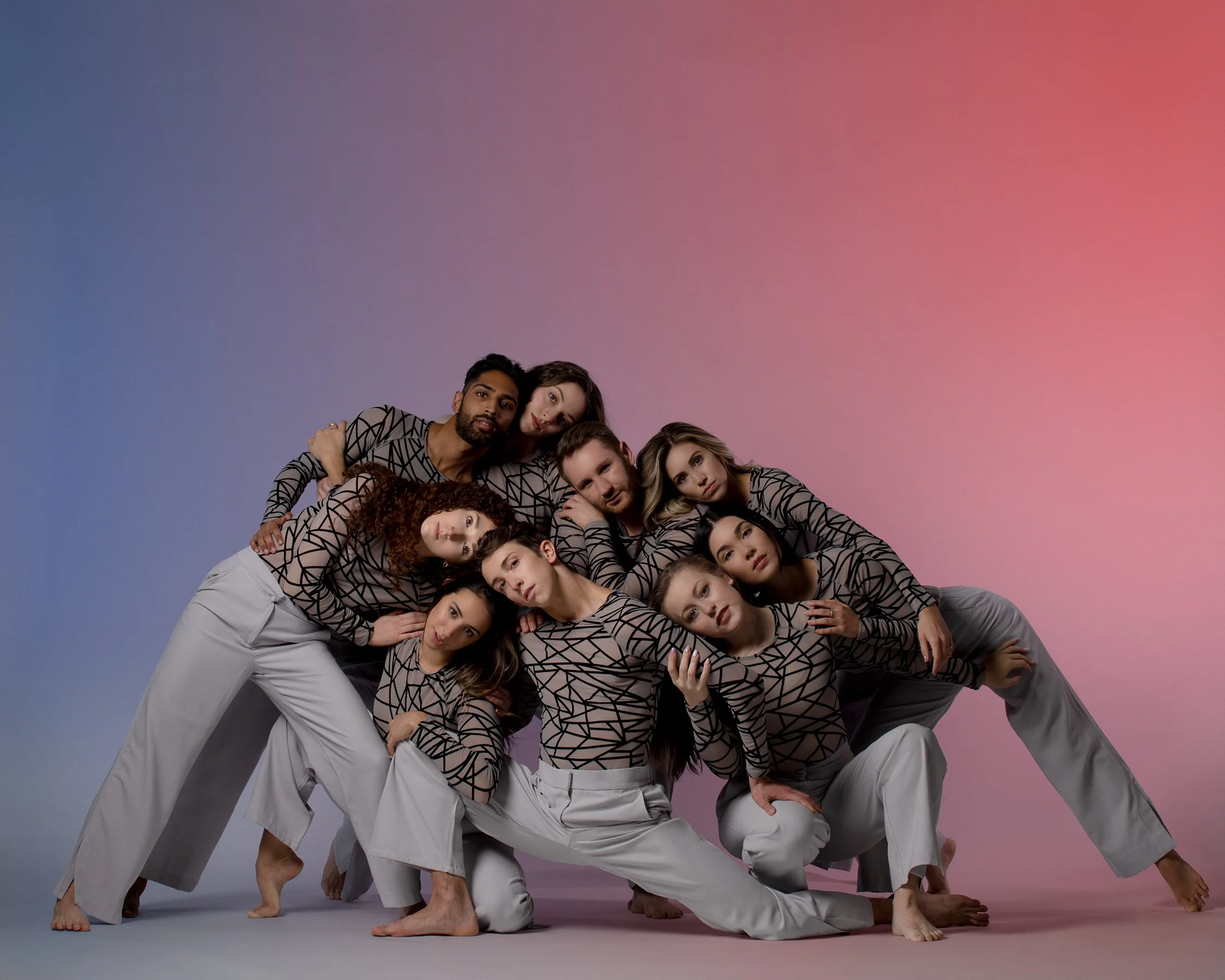 Nine Eisenhower Dance Detroit company dancers pose together during a promotional photoshoot. They are leaning on one another while wearing light gray pants and gray, mesh, fitted shirts with black geometric patterning.