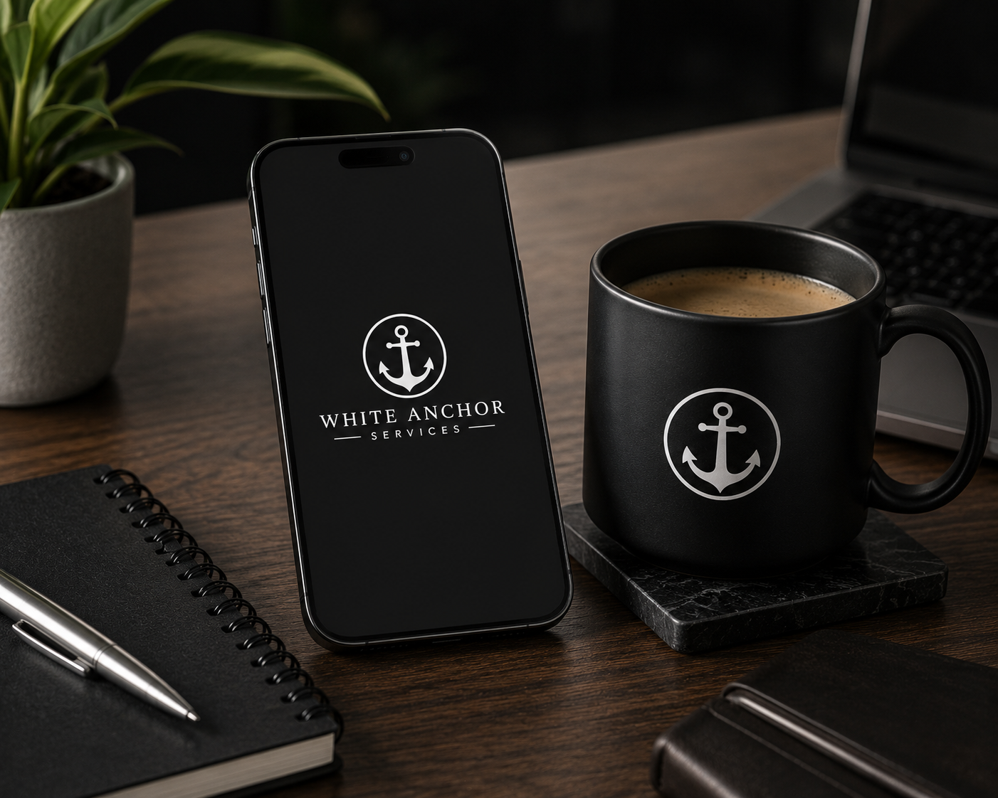 A desk scene with a smartphone displaying the logo for White Anchor Services, a black coffee mug with the same logo, a laptop, a notebook with a pen, a plant in a pot, and a closed notebook.