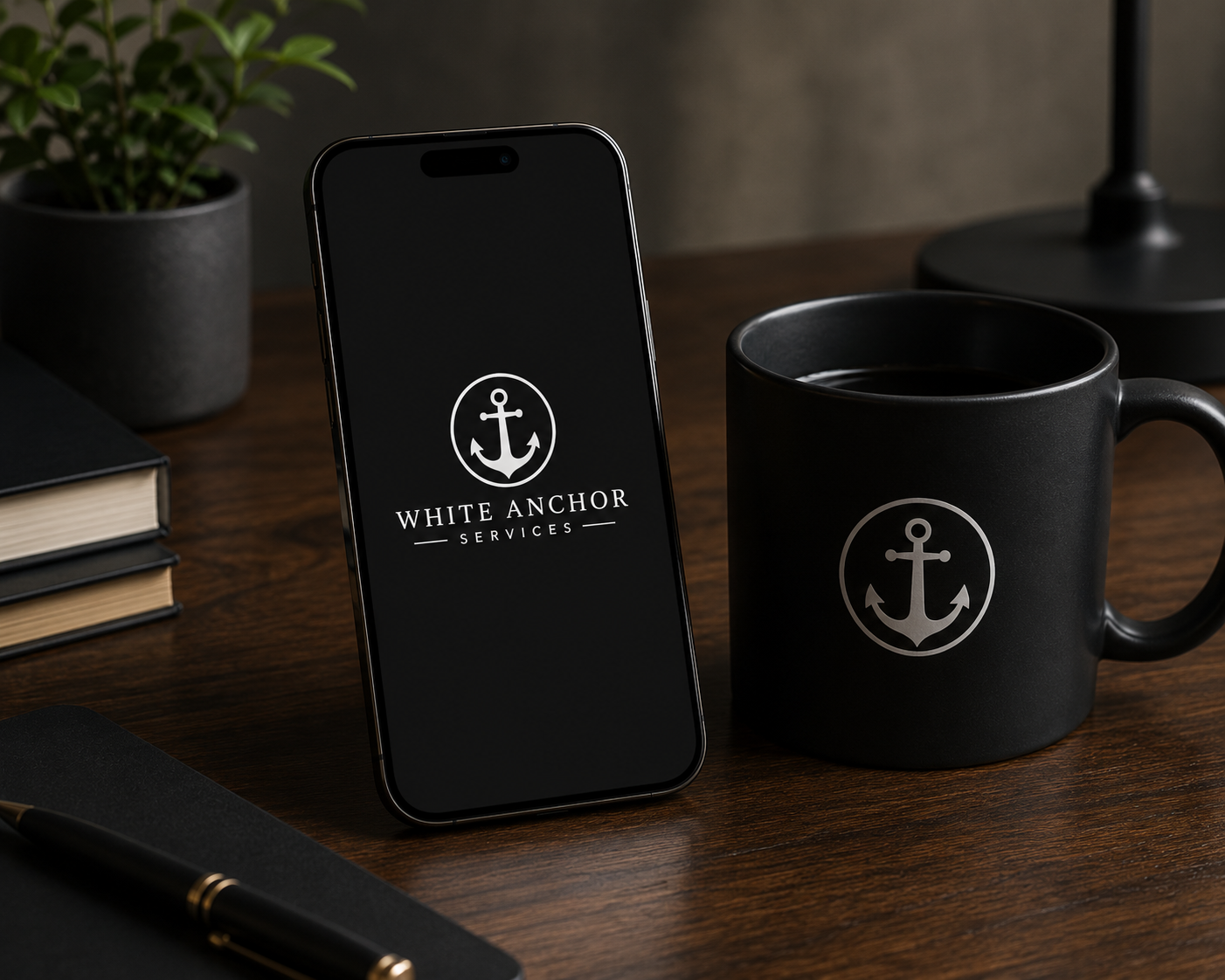 A smartphone on a wooden desk displaying the White Anchor Services logo, which is a white anchor inside a circle with the company name below. Next to the phone is a black mug with the same logo, filled with a dark beverage. There are also a closed notebook with a black cover, a black pen with gold accents, a small potted plant, and a black desk lamp in the background.