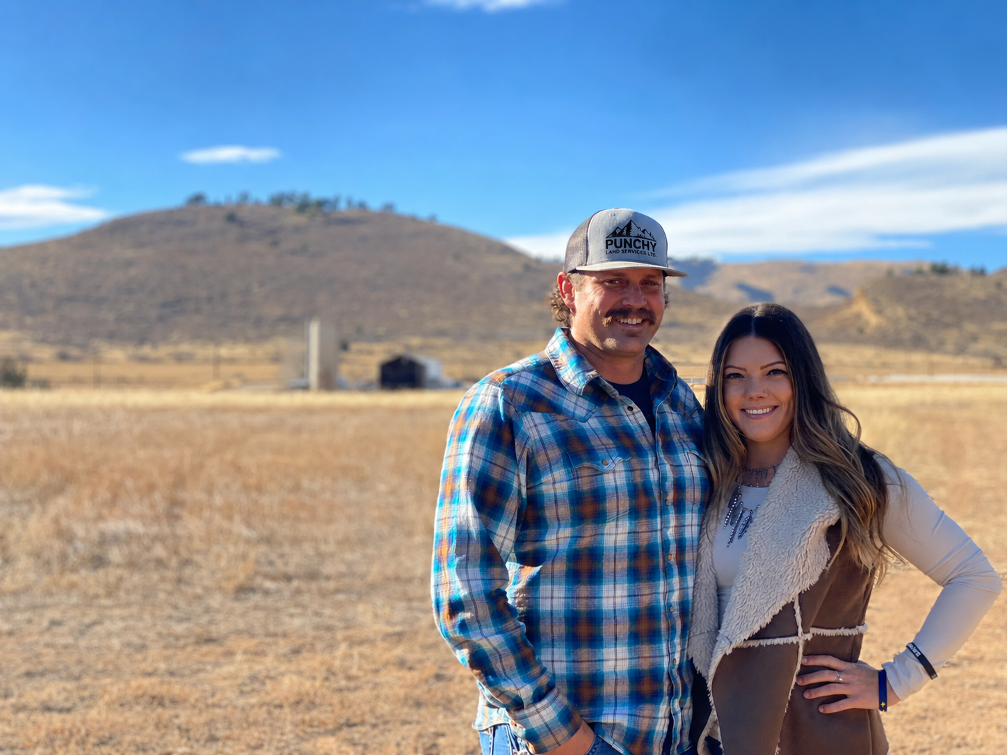 A man and woman smiling outdoors in a rural area with dry grass, hills, and a blue sky in the background.