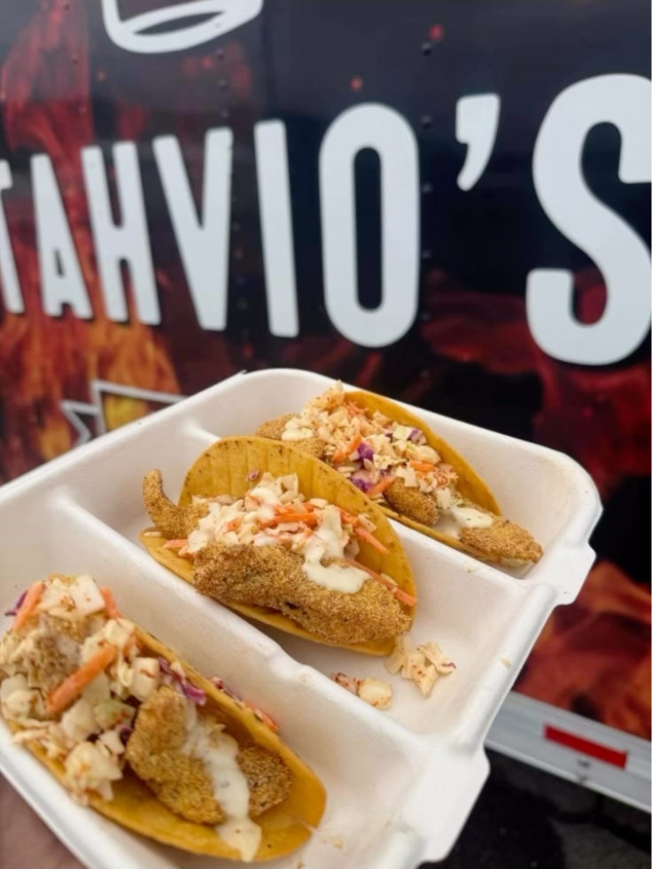 Three fish tacos in a white foam tray with shredded cabbage, sauce, and fried fish fillets on corn tortillas, against a blurred background with text.