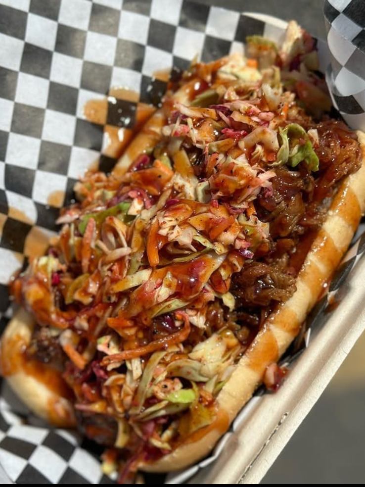 A loaded beef hot dog topped with shredded lettuce, diced tomatoes, shredded cabbage, barbecue sauce, and other condiments in a toasted bun.