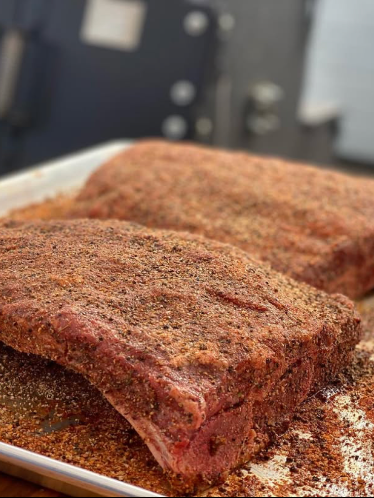 Two large seasoned slabs of raw beef brisket on a white tray, coated with a dry rub of spices, ready for smoking or cooking.