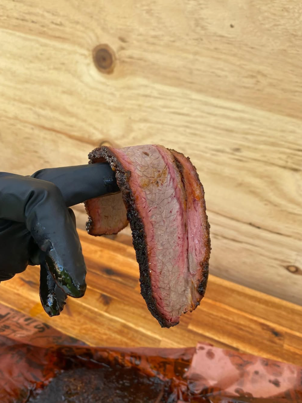 A person wearing a black glove holding a slice of smoked brisket with a dark bark, against a wooden background.