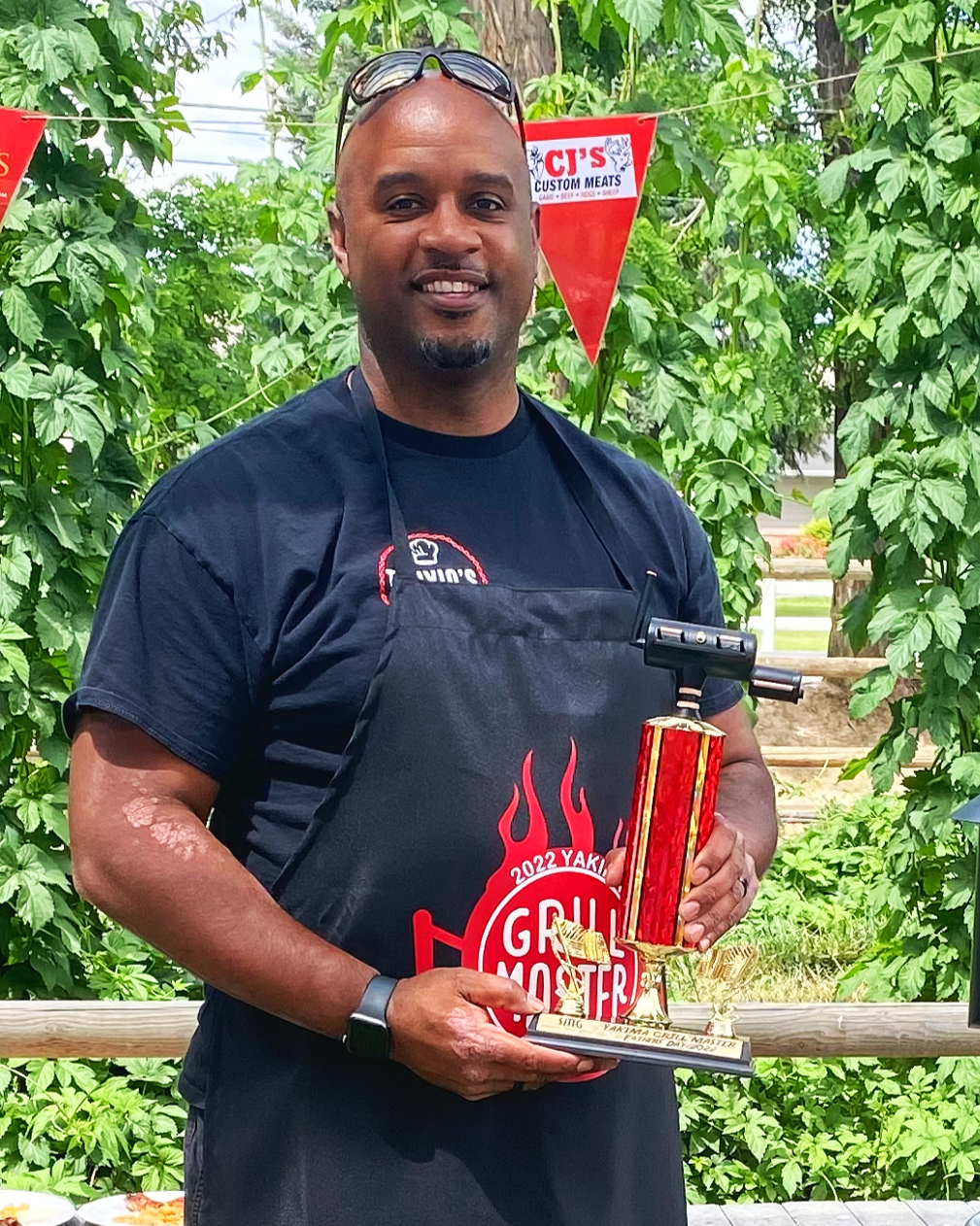 A man smiling and holding a trophy, standing outdoors with green leafy plants in the background. He is wearing a black apron with red and white text, sunglasses on his head, and a black T-shirt.