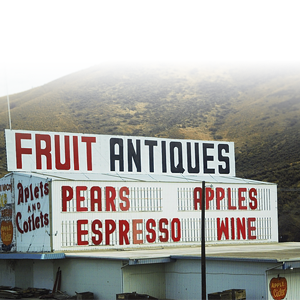 Sign advertising fruit, antiques, pears, espresso, apples, wine, and apricots in front of a hillside background.