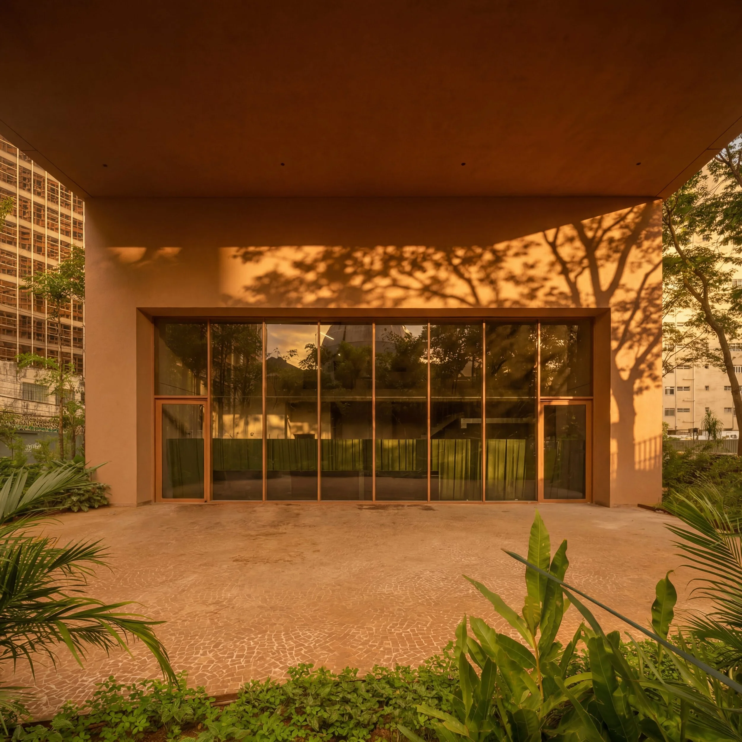 Fachada de um edifício com muitas janelas e árvores ao redor, vista ao entardecer com sombras projetadas pelo pôr do sol
