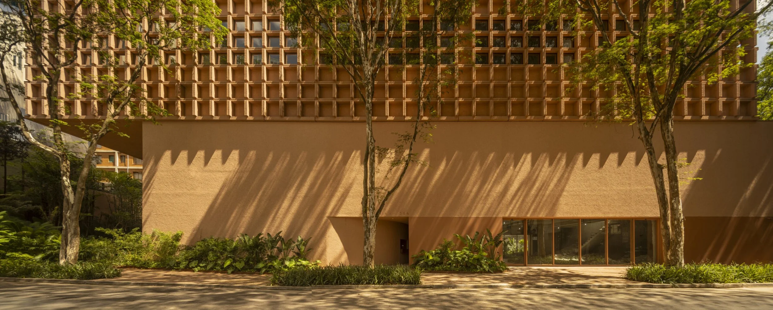 Edifício moderno com fachada de tijolos e vegetação ao redor, sombras de árvores na parede