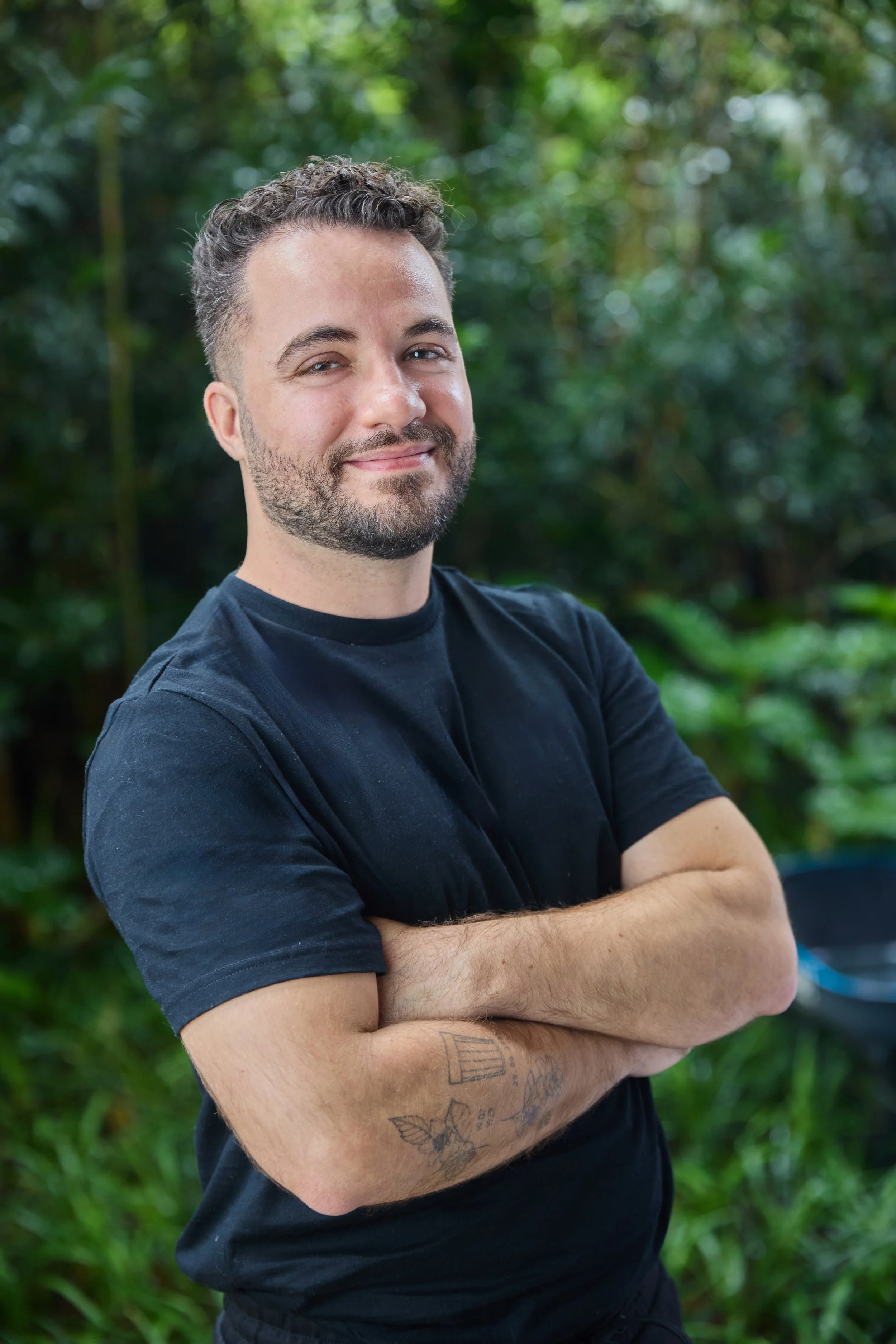 Homem de cabelo cacheado, barba, usando camiseta preta, com braços cruzados, sorrindo, ao ar livre, com fundo de vegetação verde.