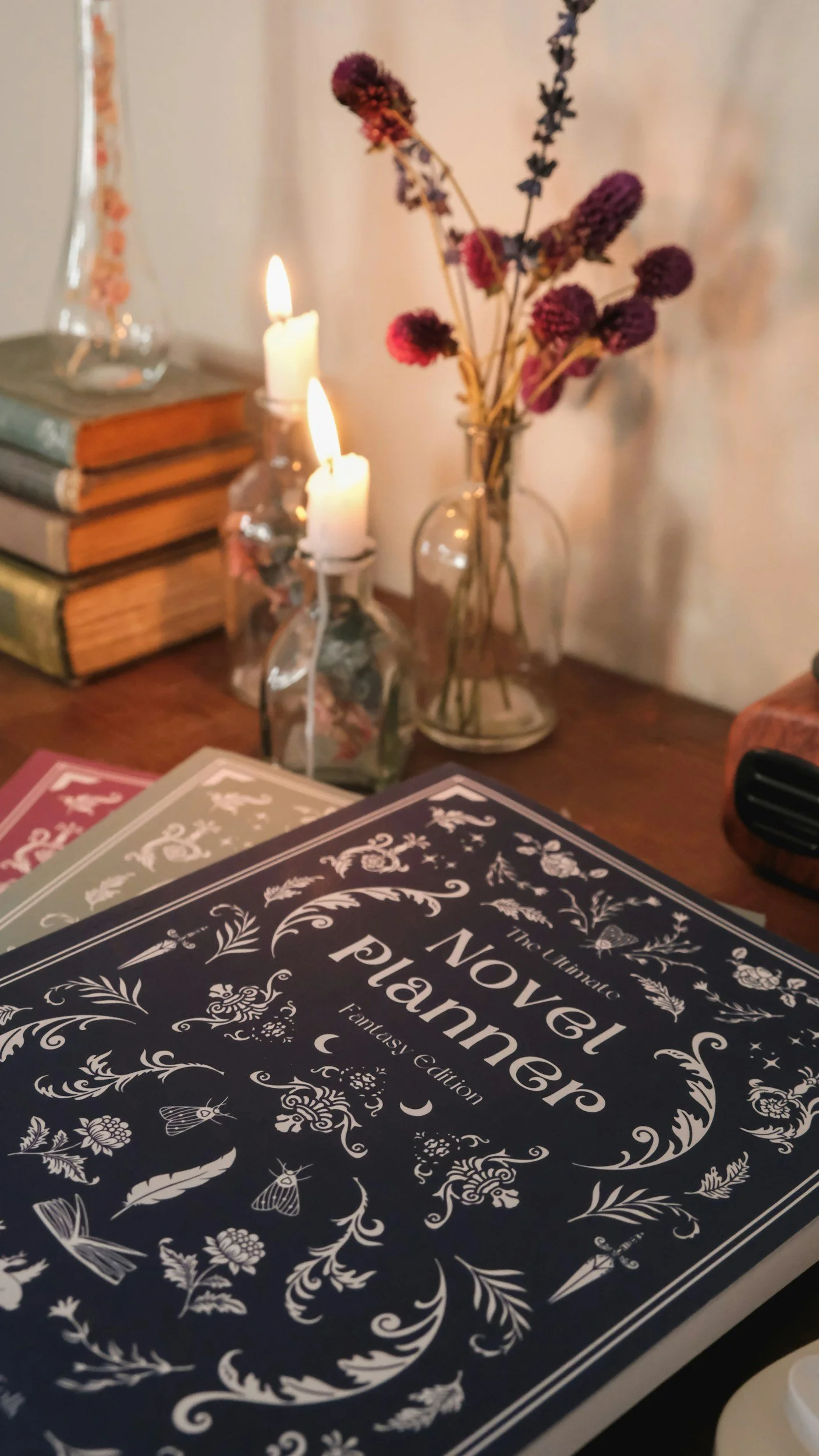 Close-up of a black and white book titled "The Novel Planner: A Fantasy Edition" on a table, with a background of books, candles, and a vase with dried flowers.