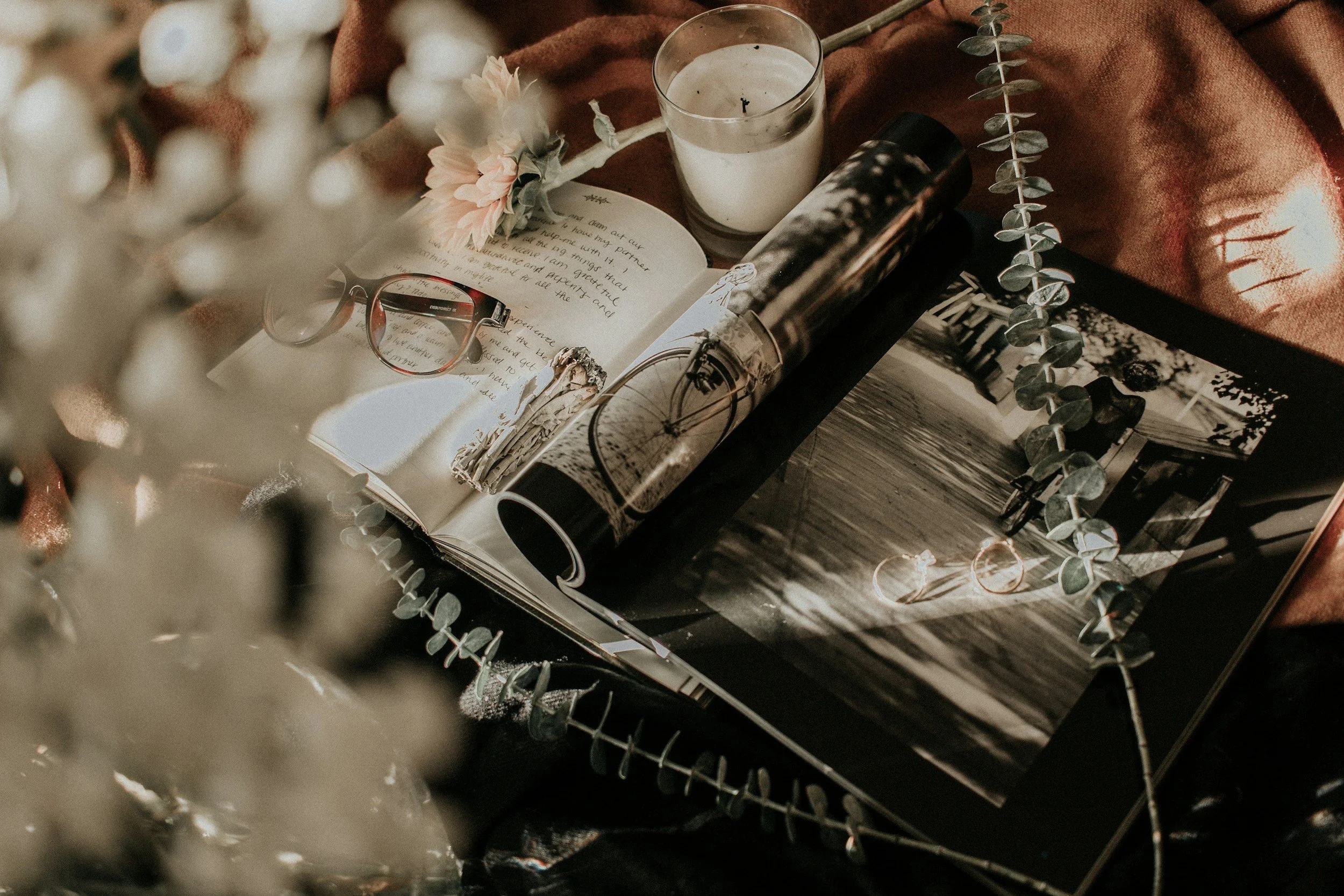 A flat lay of a workspace with a pair of reading glasses, a candle, a magazine with bicycles on the cover, an open letter, a pink flower, and a string of eucalyptus leaves, with a brown cloth in the background.