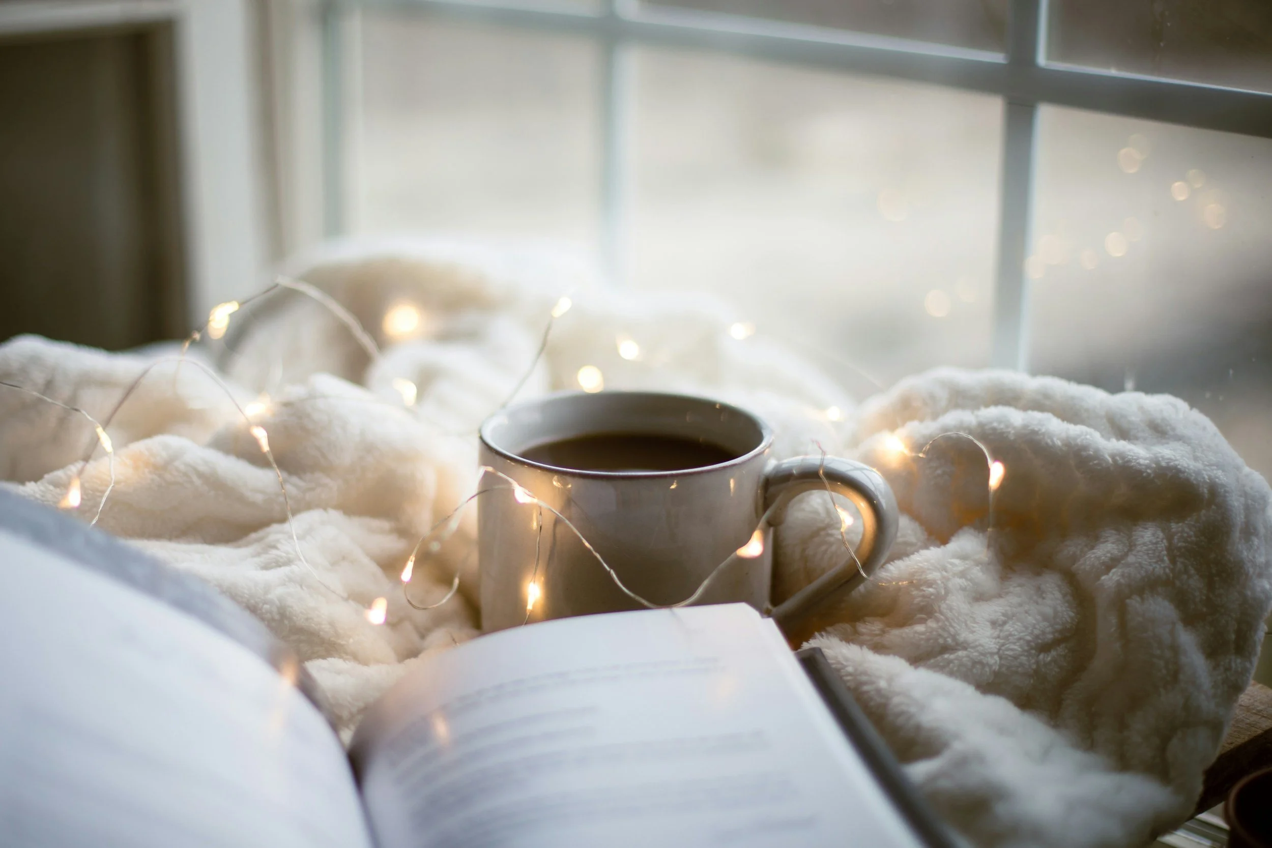 A white mug filled with coffee, placed on a fluffy white blanket with fairy lights around it, near a window with daylight.