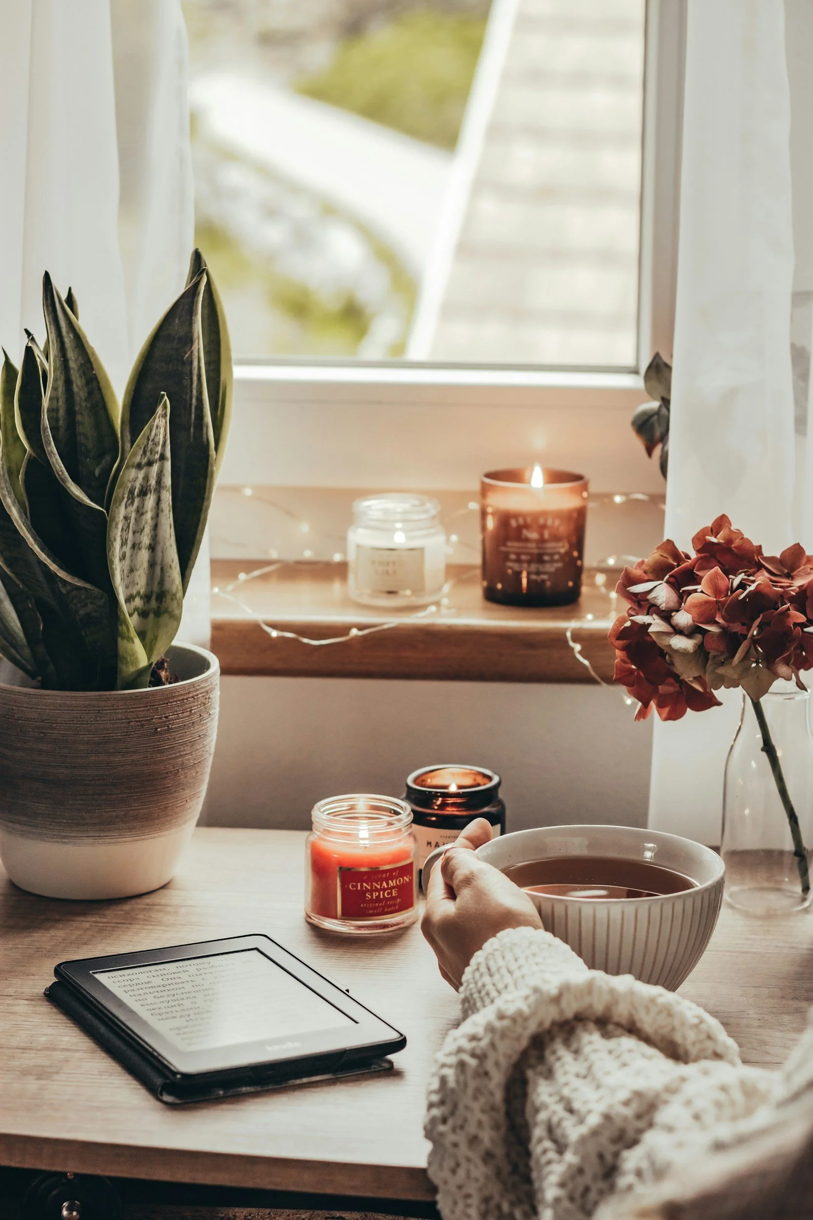 A person wearing a white knitted sweater is holding a bowl of tea in front of a window. On the table, there are candles, an e-reader with text, a potted snake plant, a pinkish hydrangea flower, and a tablet. The scene is cozy and well-lit by natural light.
