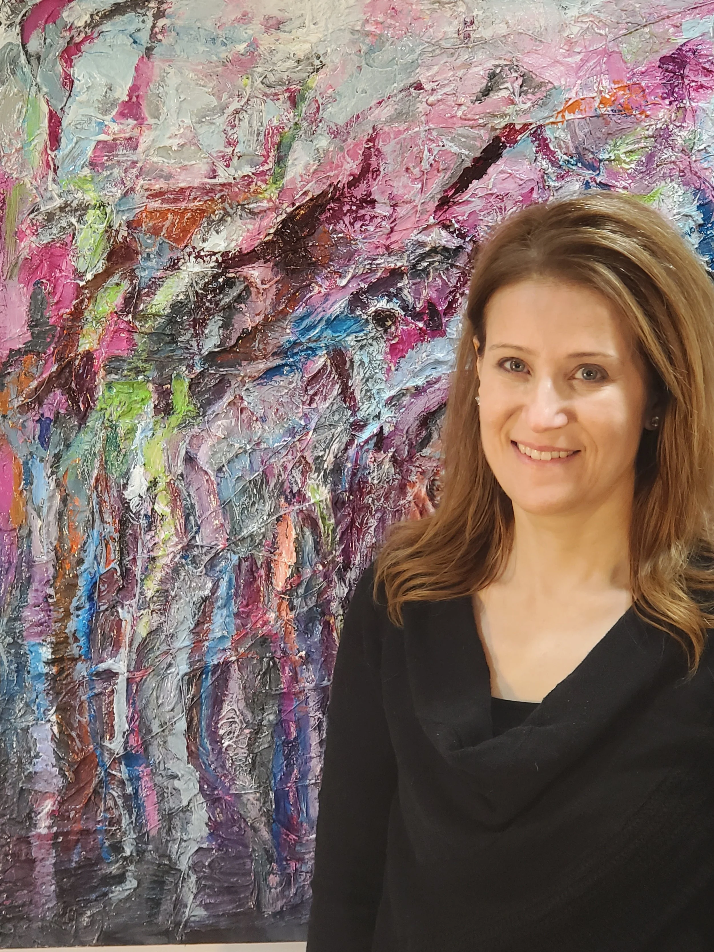 A smiling woman with shoulder-length brown hair stands next to an abstract colorful painting with pink, white, black, green, blue, and orange paint on a wall.