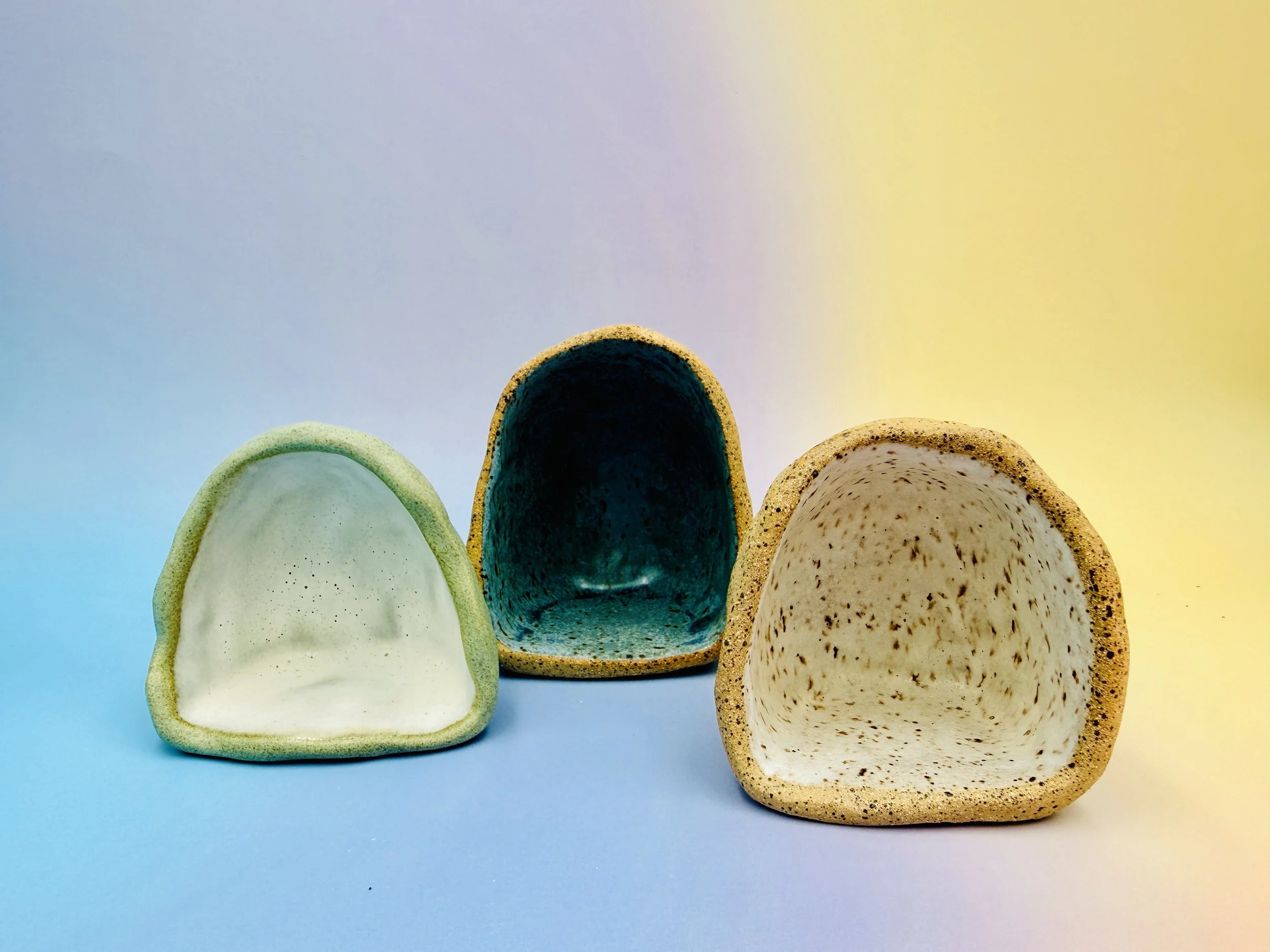Three dome-shaped ceramic sculptures with speckled interiors in light green, teal, and beige sit on a blue-yellow gradient background, creating a calming vibe.