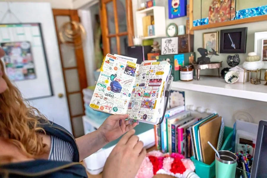 A woman with curly red hair holding an open colorful sticker or scrapbook book in a cluttered home office or art space, with shelves filled with books, artwork, and decorative objects.