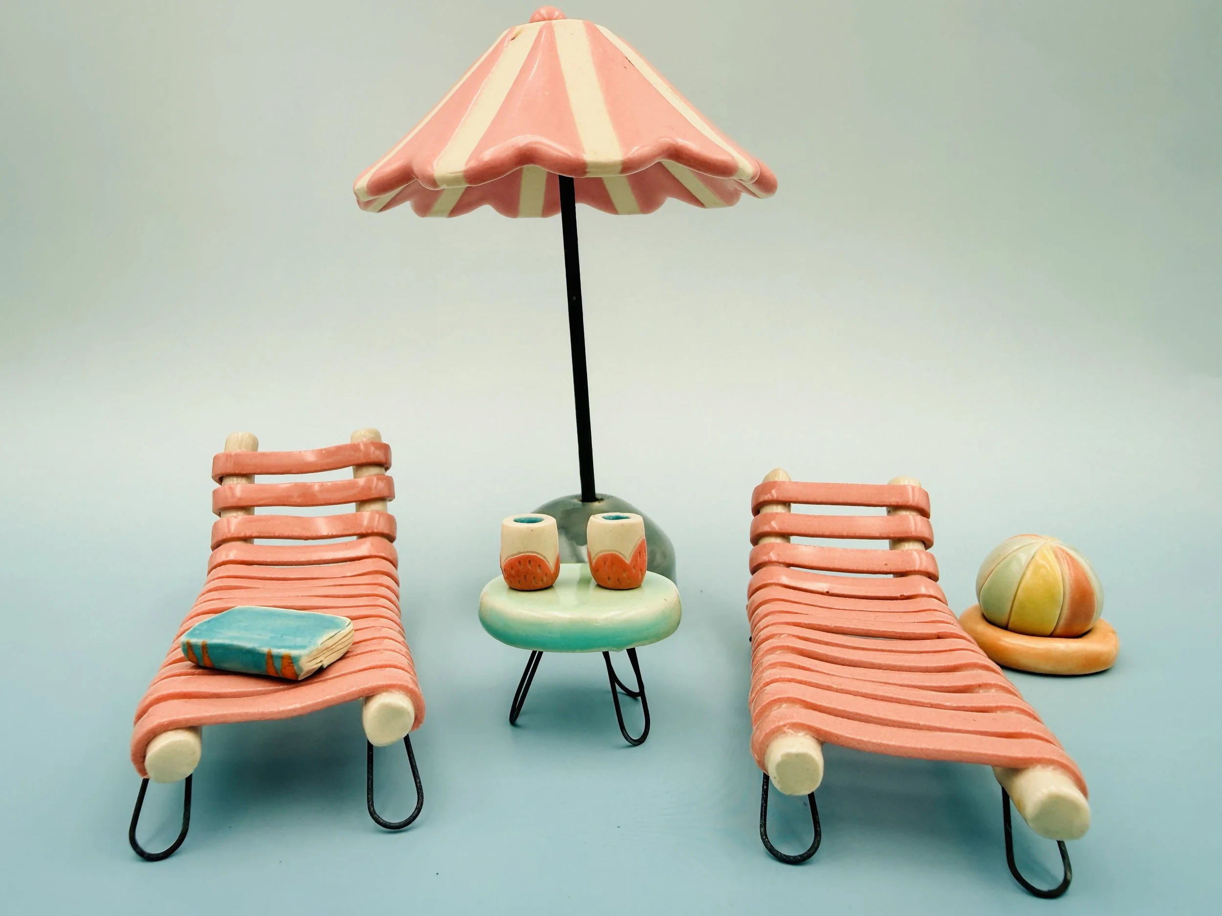 The image features a small, whimsical beach scene created with ceramic figurines. In the foreground, there are two brightly colored lounge chairs positioned on either side of a small round table. The chairs have a striped pattern in light pink and cr