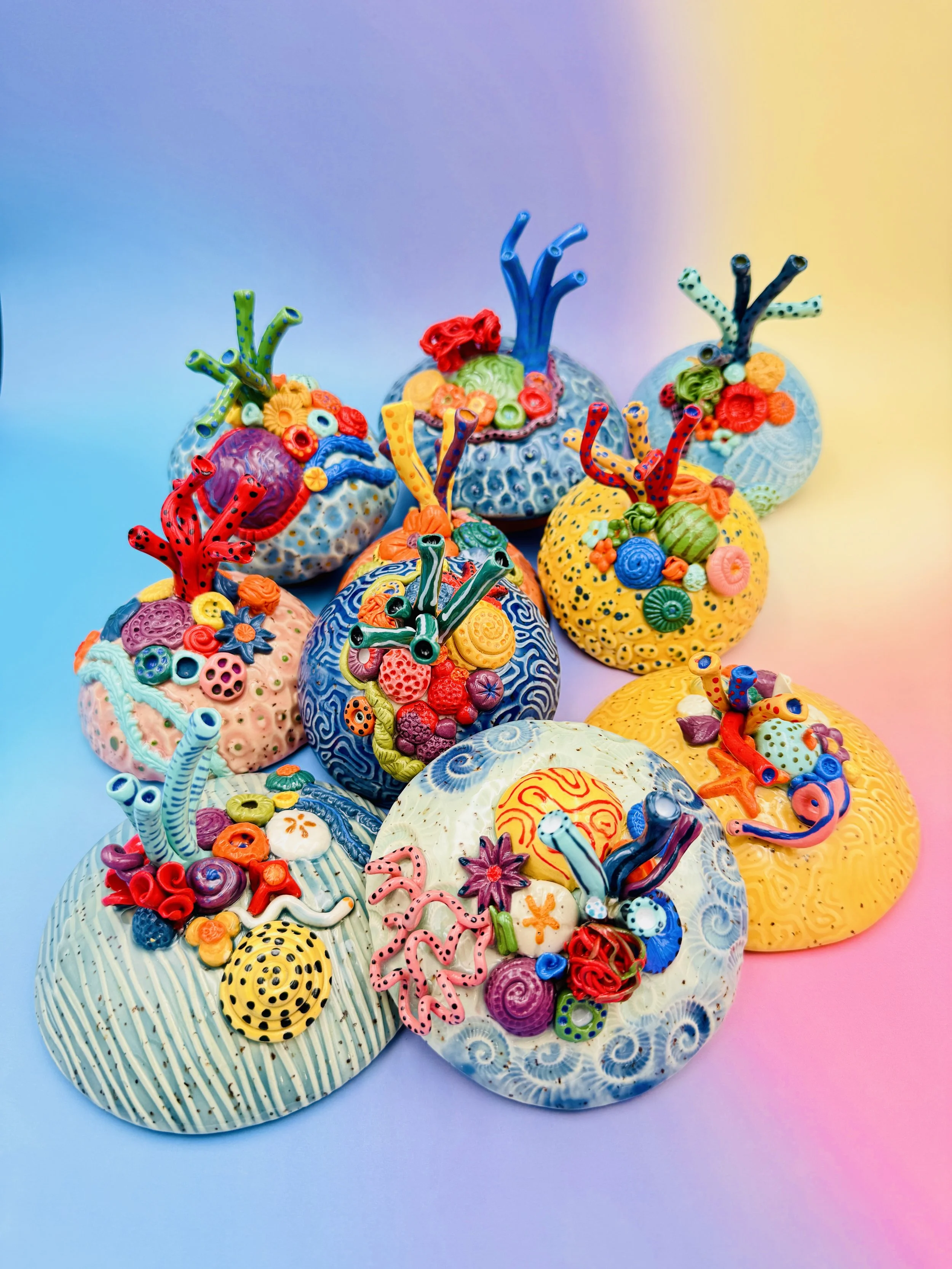 Colorful, textured clay sculptures resembling coral reefs arranged on a gradient background. Each piece has vibrant, intricate details, evoking a sense of ocean life.